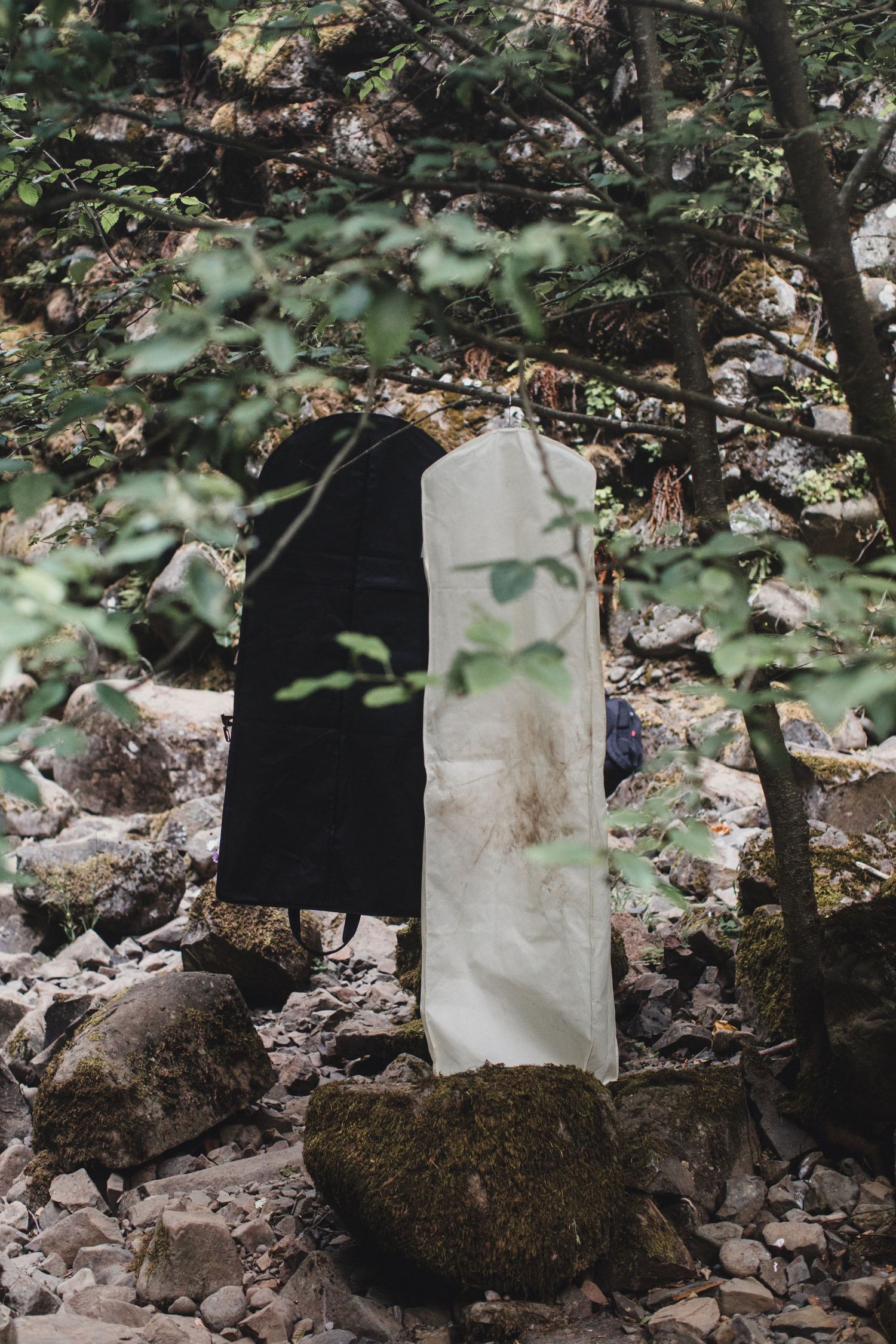 Wedding dress and suit in garment bags hanging on a mossy rock in a forest setting.