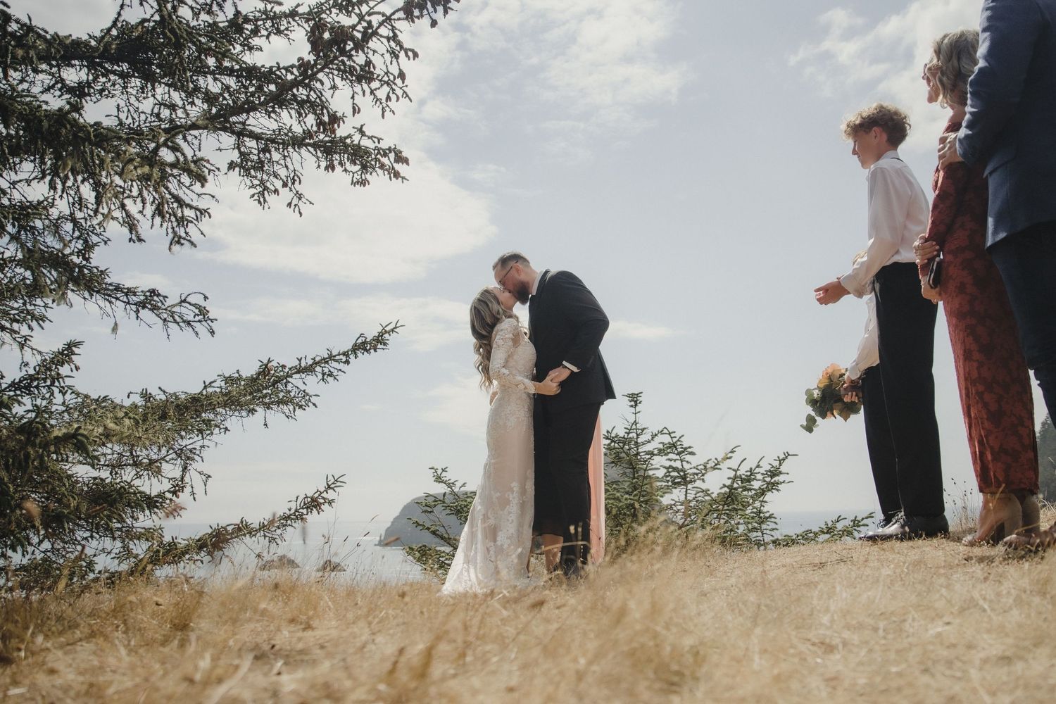 Bride and groom share their first kiss during an outdoor wedding ceremony beneath a tree with guests watching.