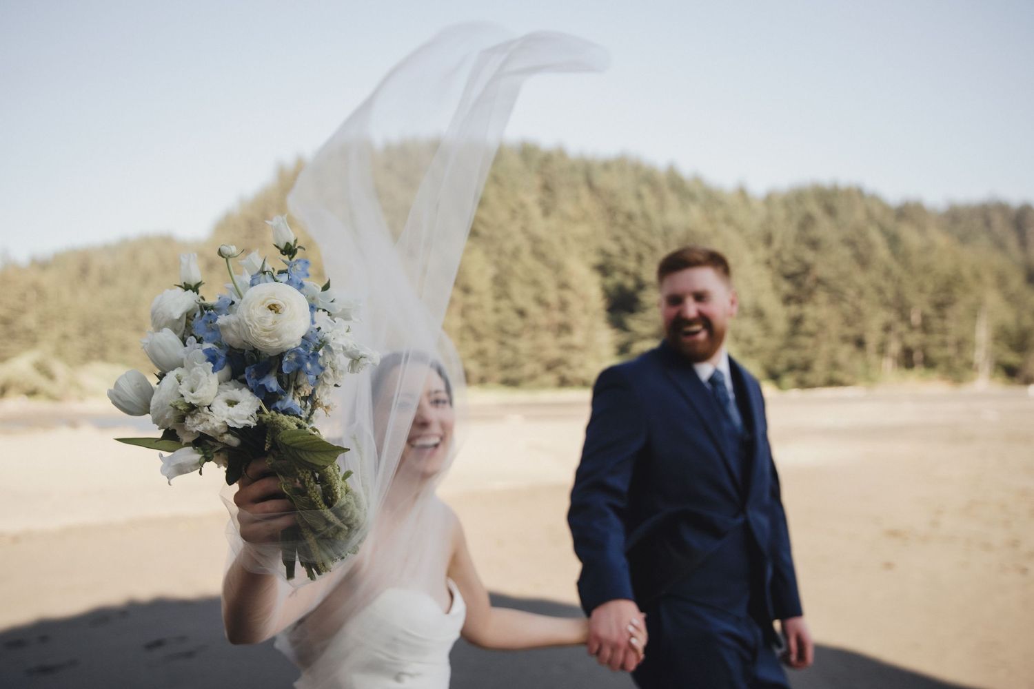 Bride laughing as veil blows in wind holding blue and white bouquet with groom on beach.