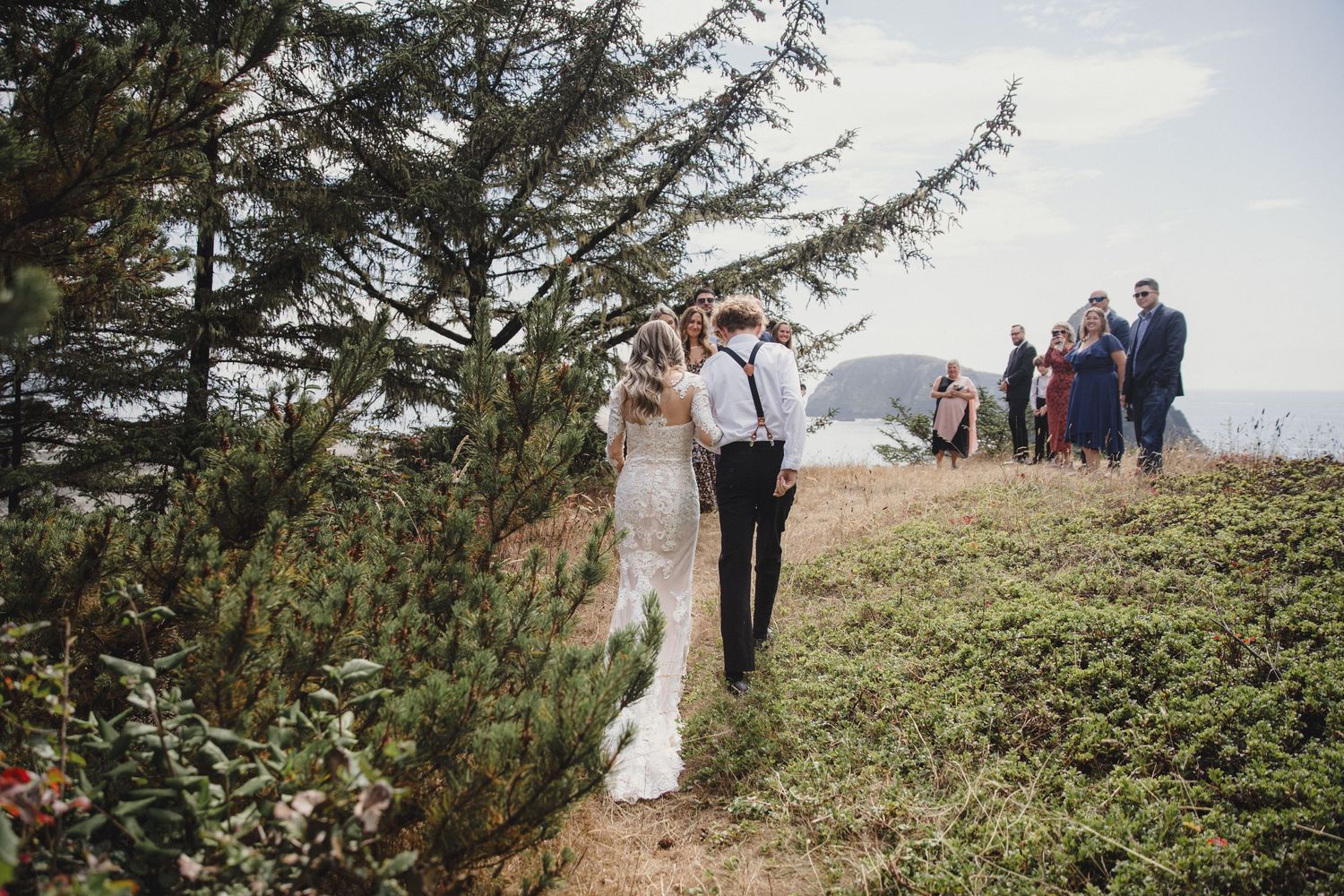 Newlywed couple walks along a scenic coastal hillside path with guests and ocean views in the background.