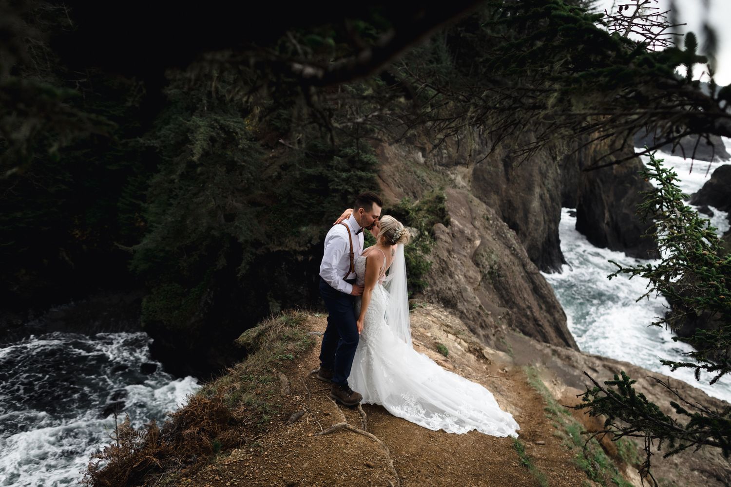 A romantic moment captured on a cliff edge with dramatic ocean waves and evergreens in the background.