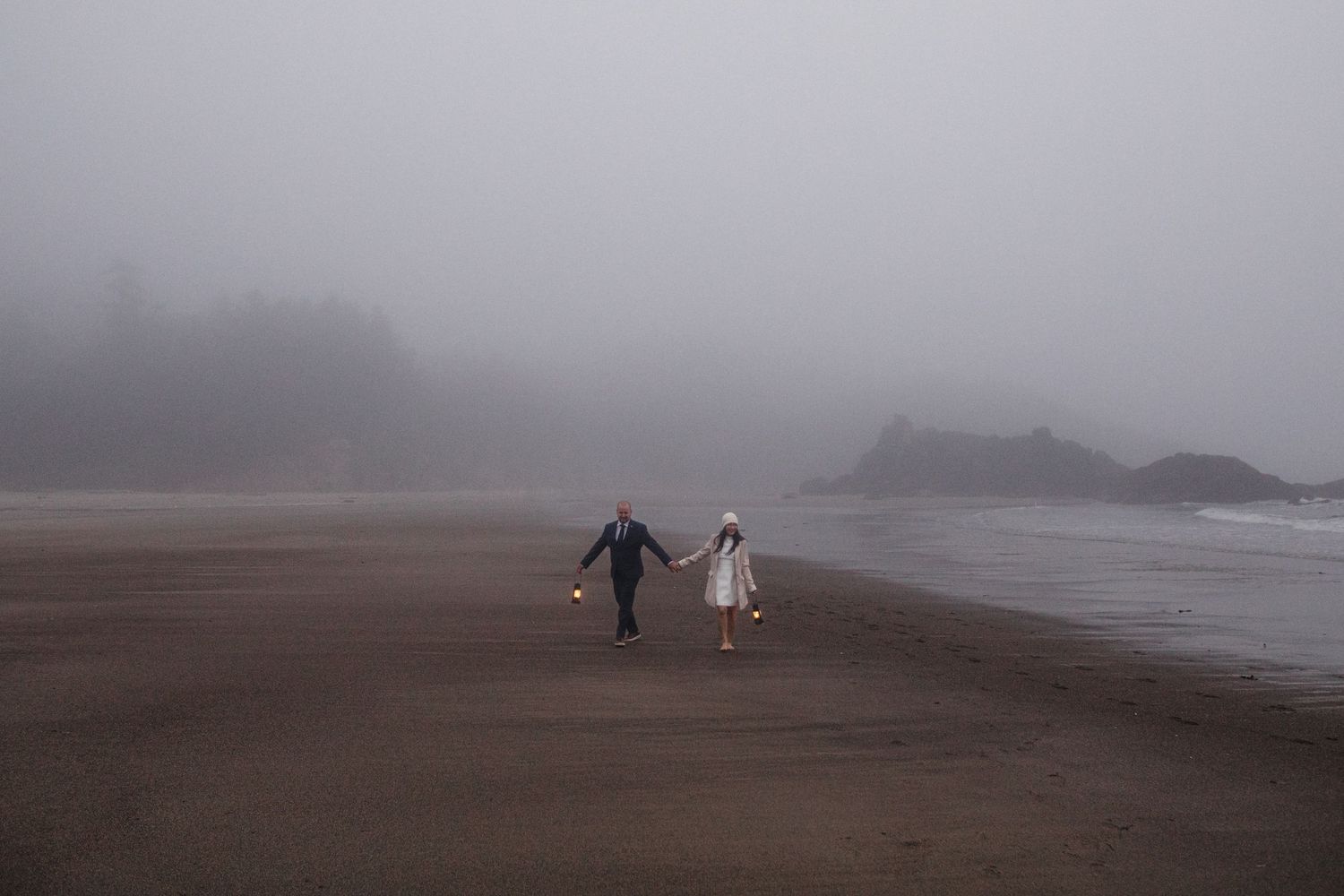 Two people walking hand in hand on a foggy beach with mountains visible in the misty distance.