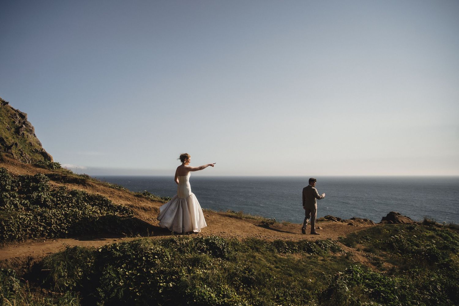 Bride in white gown and groom stand on coastal cliffside trail with ocean and mountain views on wedding day.