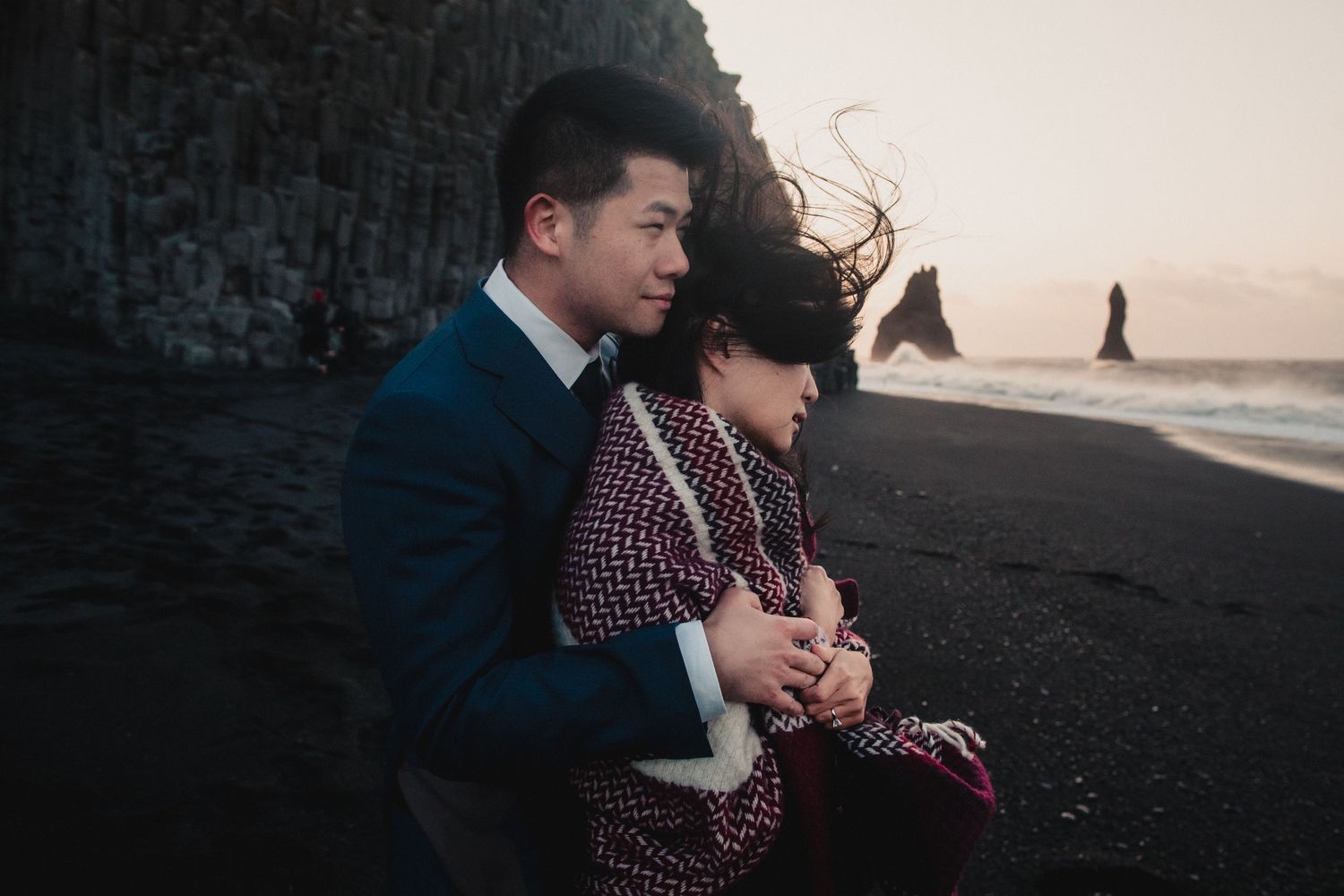 A tender embrace on a black sand beach with dramatic coastal cliffs in the background.
