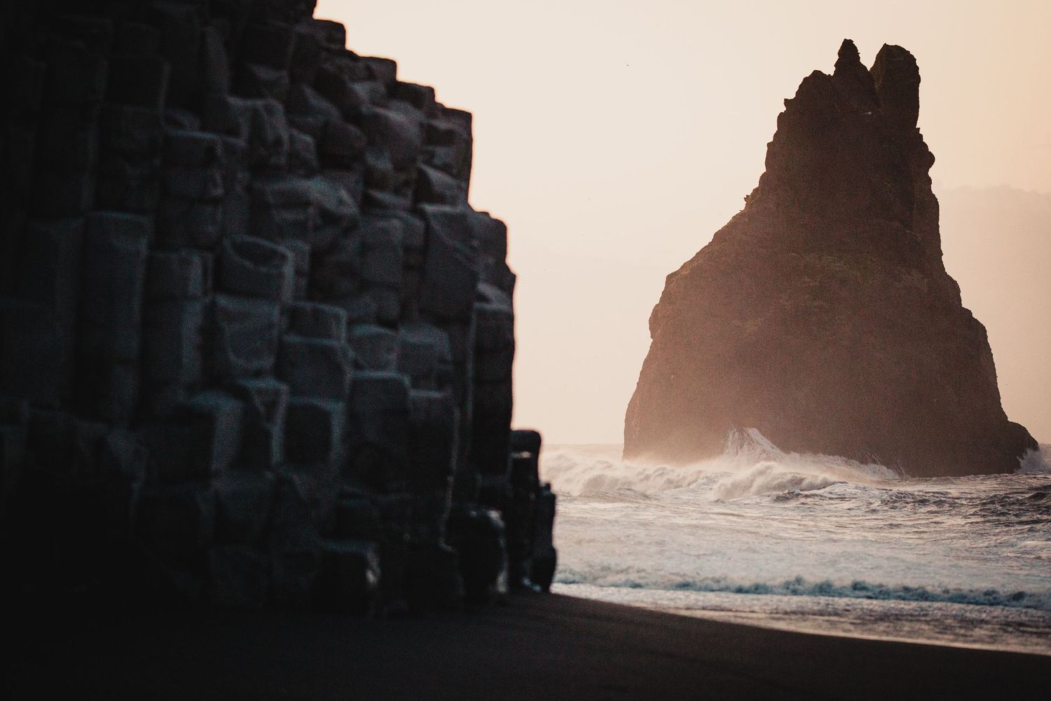 Geometric basalt cliff formations frame a dramatic sea stack on a misty beach.