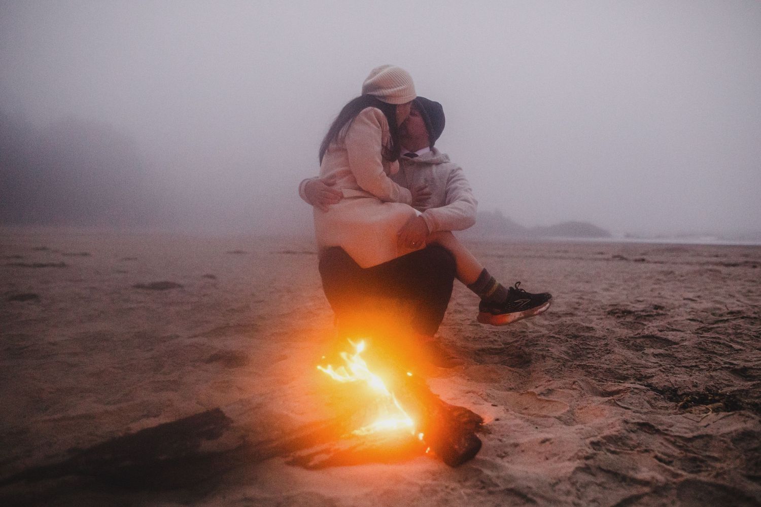 Couple embracing by a small campfire on a foggy beach, creating a romantic and moody atmosphere.