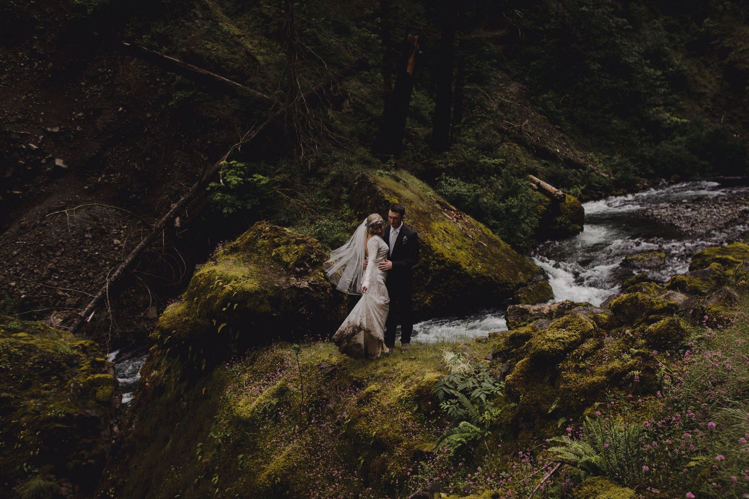 Wedding couple poses dramatically on moss-covered rocks near a rushing stream.