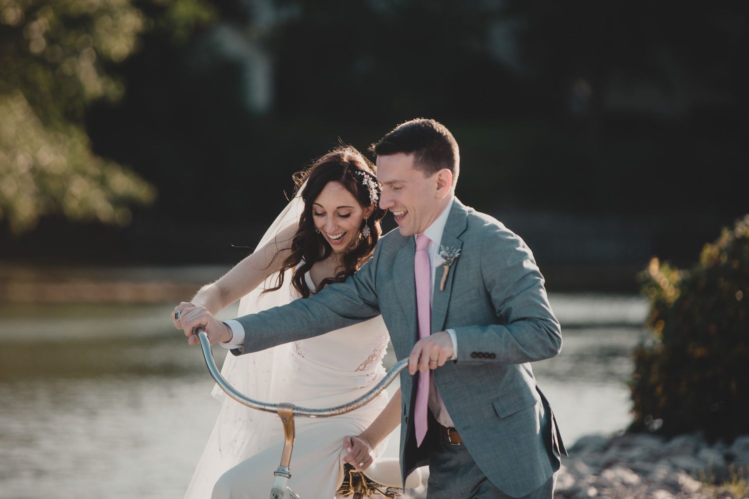 Bride and groom laughing together while riding a vintage bicycle near a sunlit river on their wedding day.