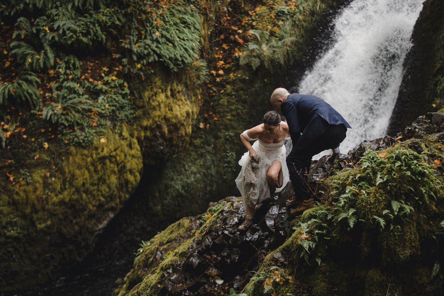 Bride and groom in wedding attire climbing mossy rocks beside a lush waterfall in a forest setting.