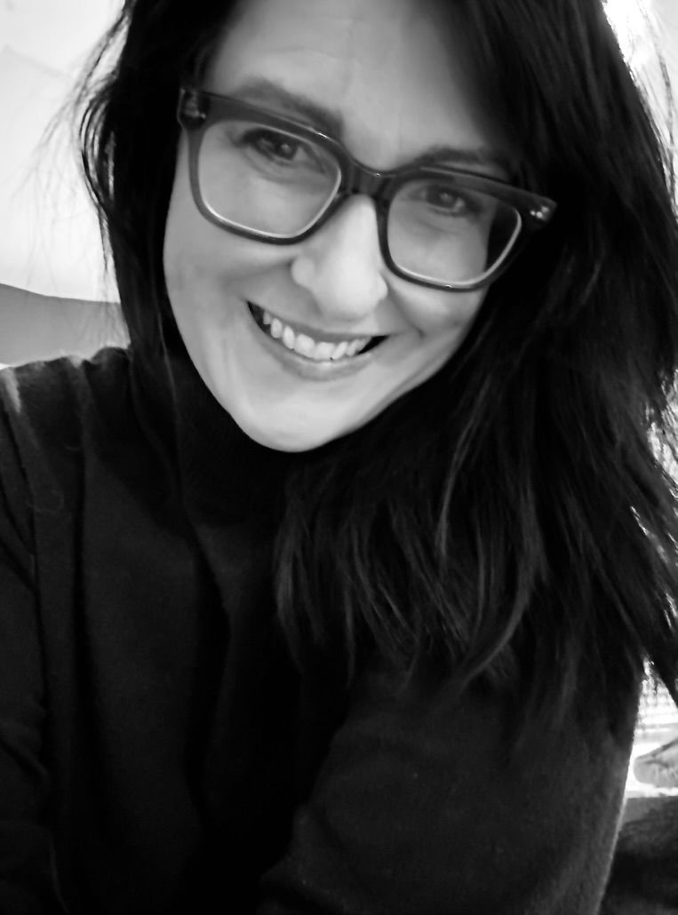 Black and white selfie of a smiling woman with dark hair and glasses wearing a turtleneck sweater.