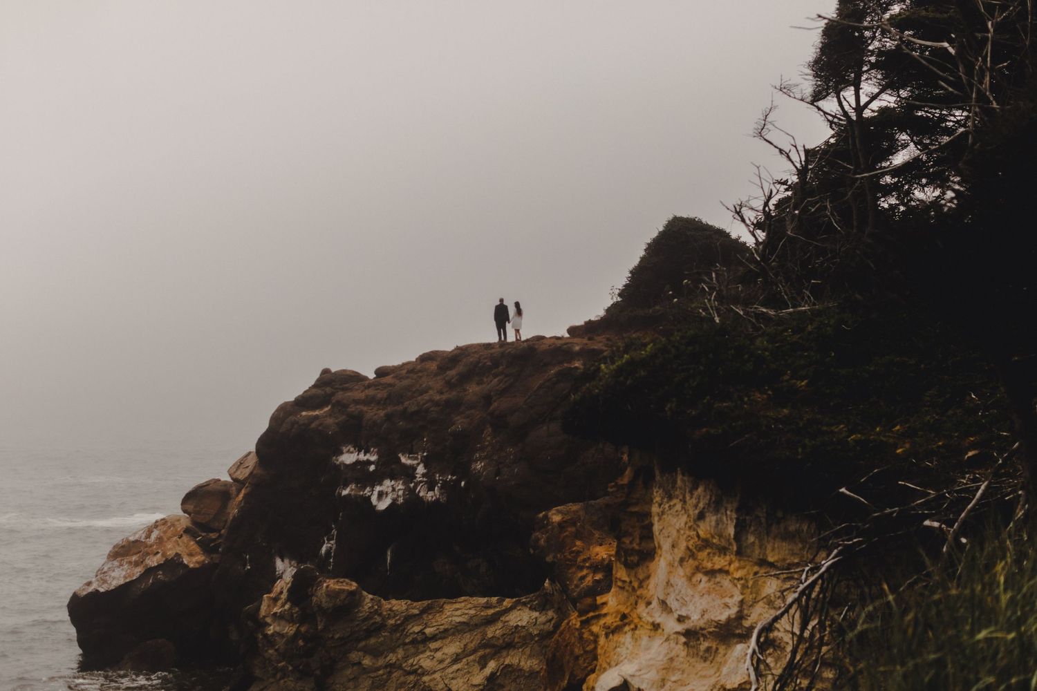 Couple standing on a dramatic coastal cliff overlooking a misty ocean on a foggy day.