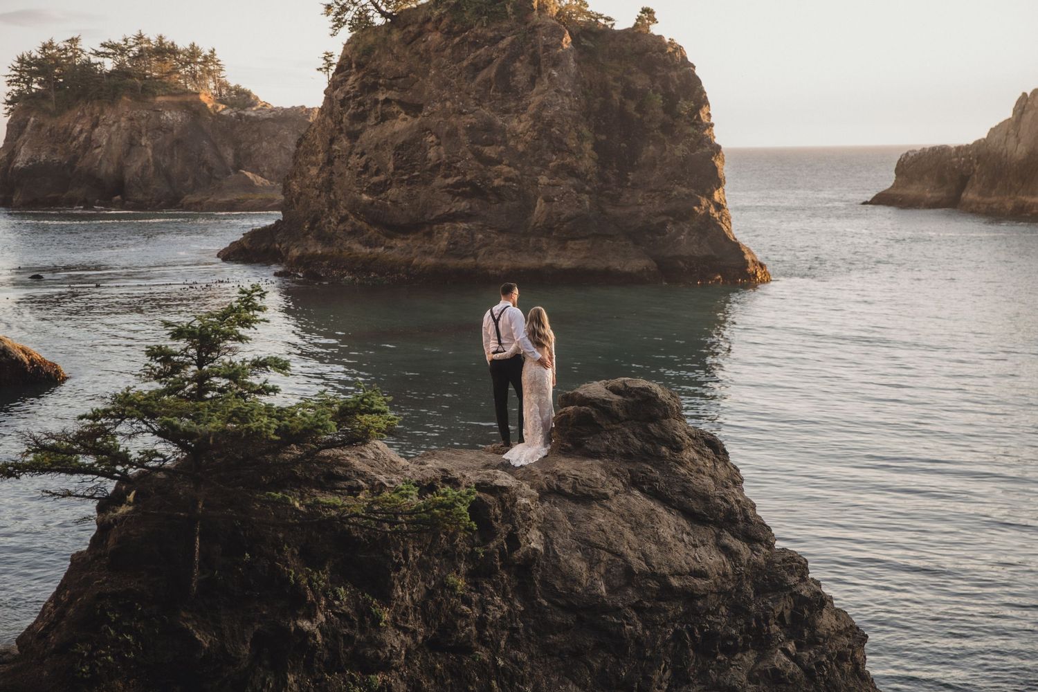 Bride and groom stand on a dramatic rocky coastal overlook surrounded by sea stacks and ocean.