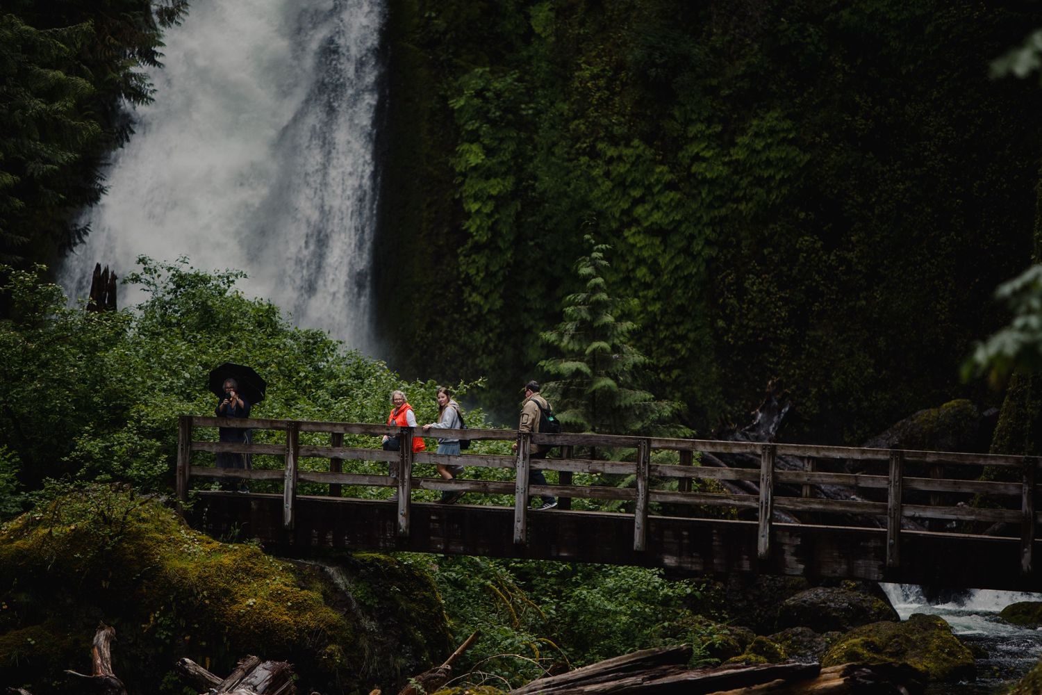 Visitors pose on wooden bridge with dramatic waterfall and forest scenery in background.