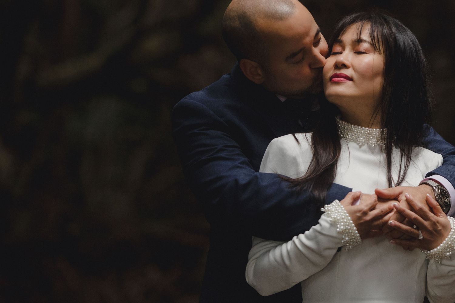 Groom tenderly kisses bride on the cheek while embracing her from behind in a dark forest.
