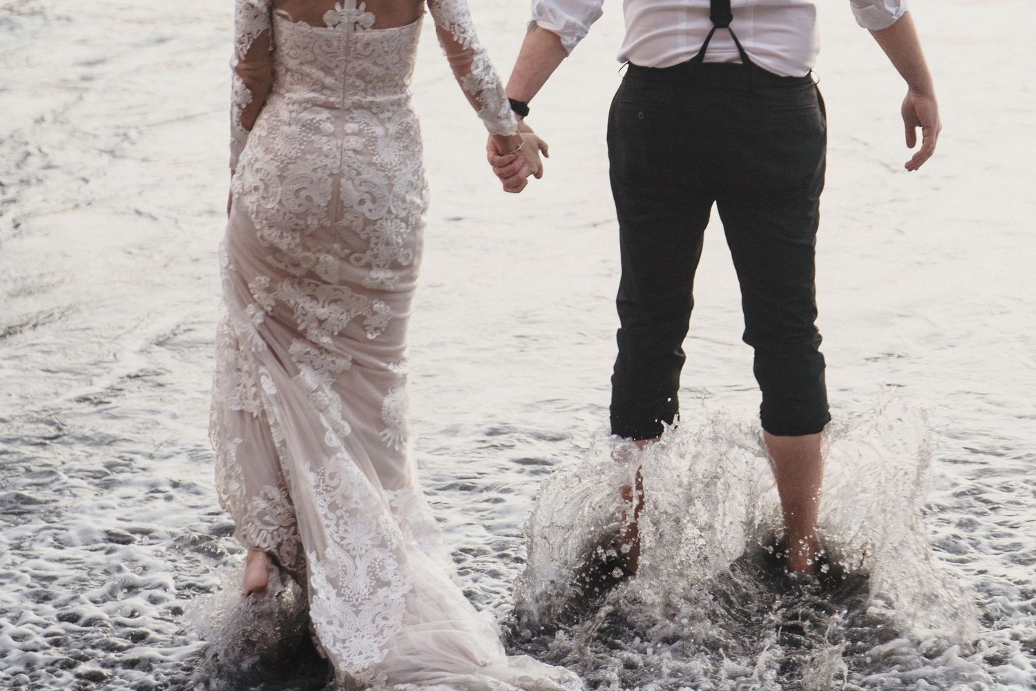 Bride in lace gown and groom in suspenders hold hands while waves splash around their feet.