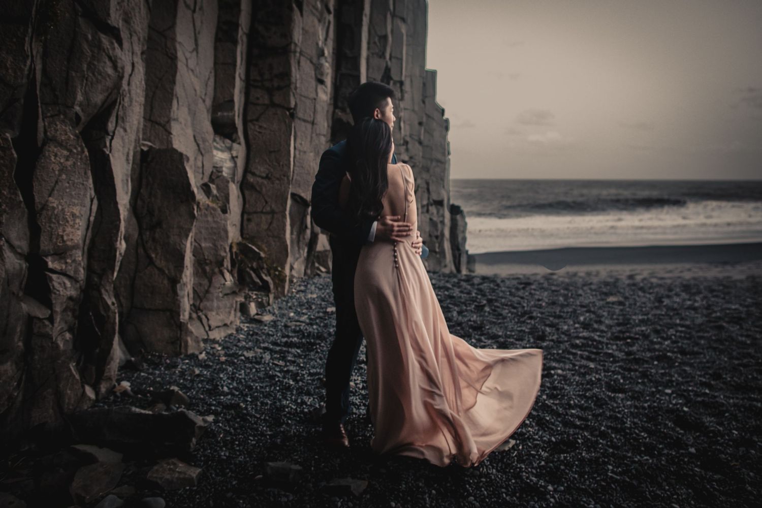 A couple poses against dramatic black basalt cliffs on a rocky beach at dusk.