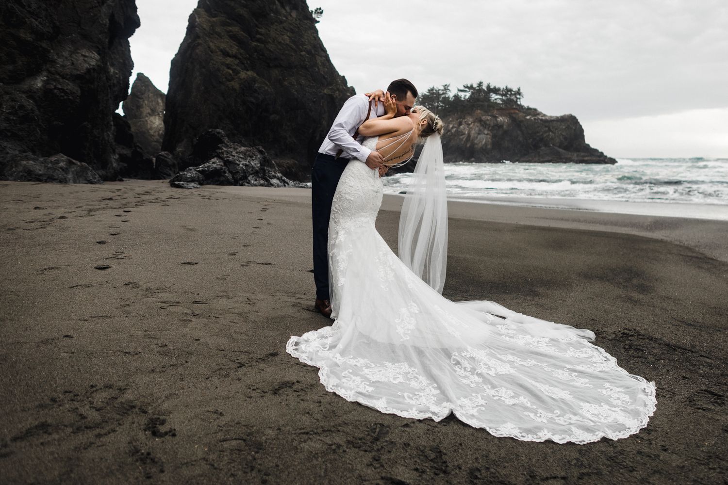 A dramatic wedding portrait on a dark sandy beach with towering cliffs and ocean waves.