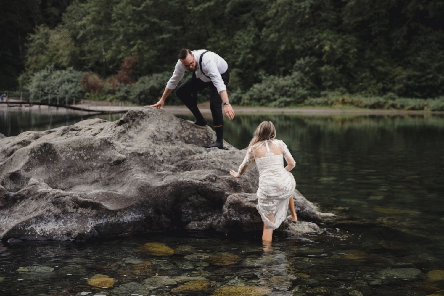Groom helps bride navigate large boulder at waters edge during adventurous outdoor elopement session.