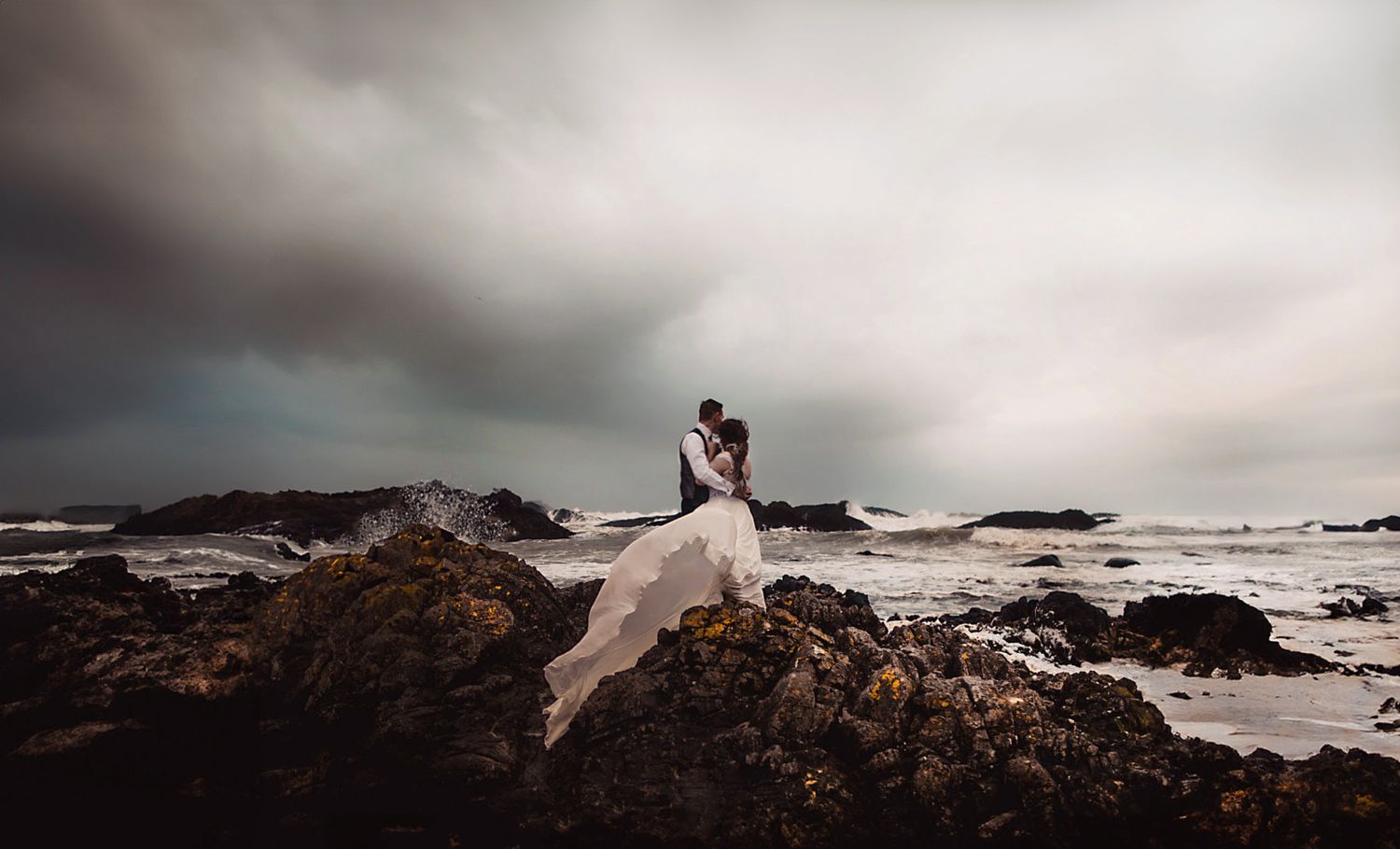 A dramatic coastal wedding photo with crashing waves and rocky shoreline.