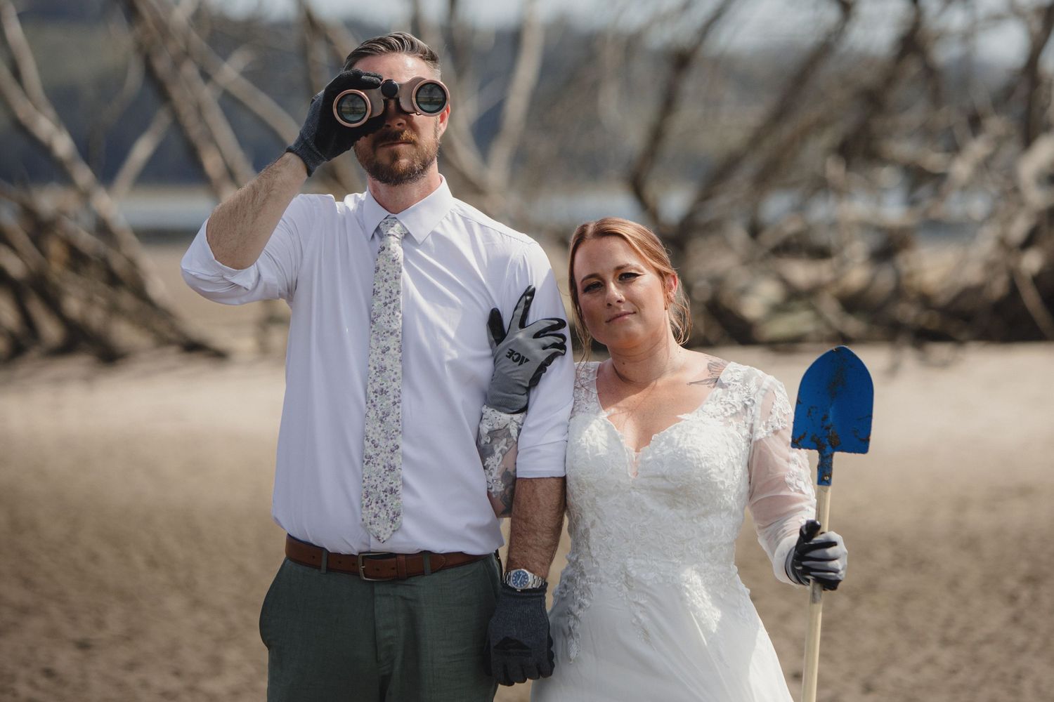 Bride in lace gown and groom in tie holding binoculars and shovel pose on sandy beach for quirky wedding photo.