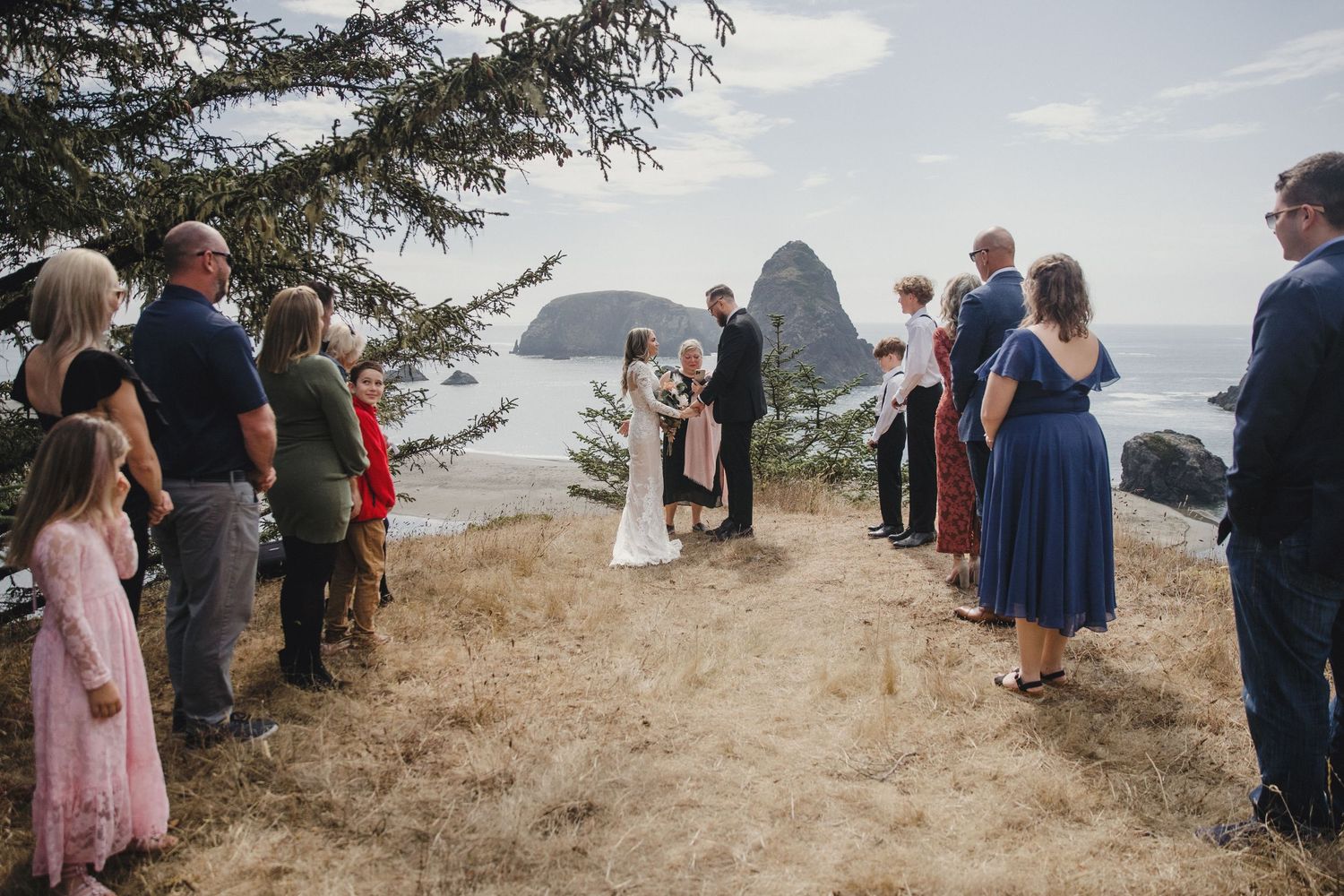 Outdoor wedding ceremony on a coastal cliff with ocean and rock formations in the background, guests gathered around the couple.