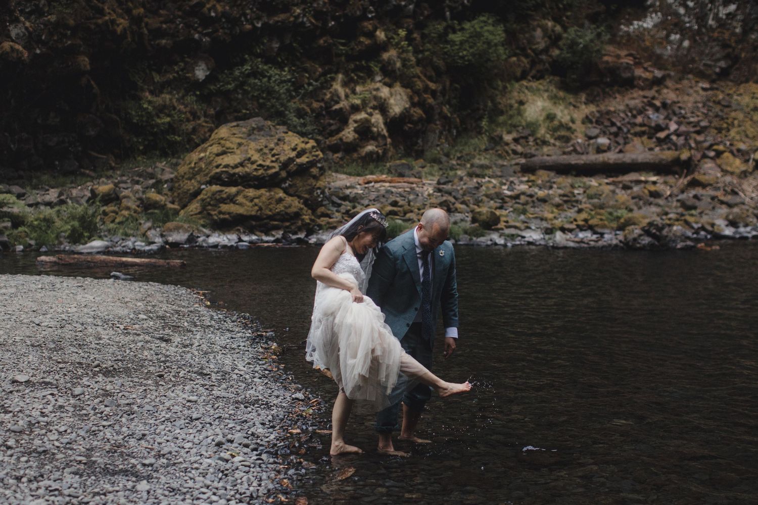 Groom helping bride lift her wedding dress while wading in a rocky stream.