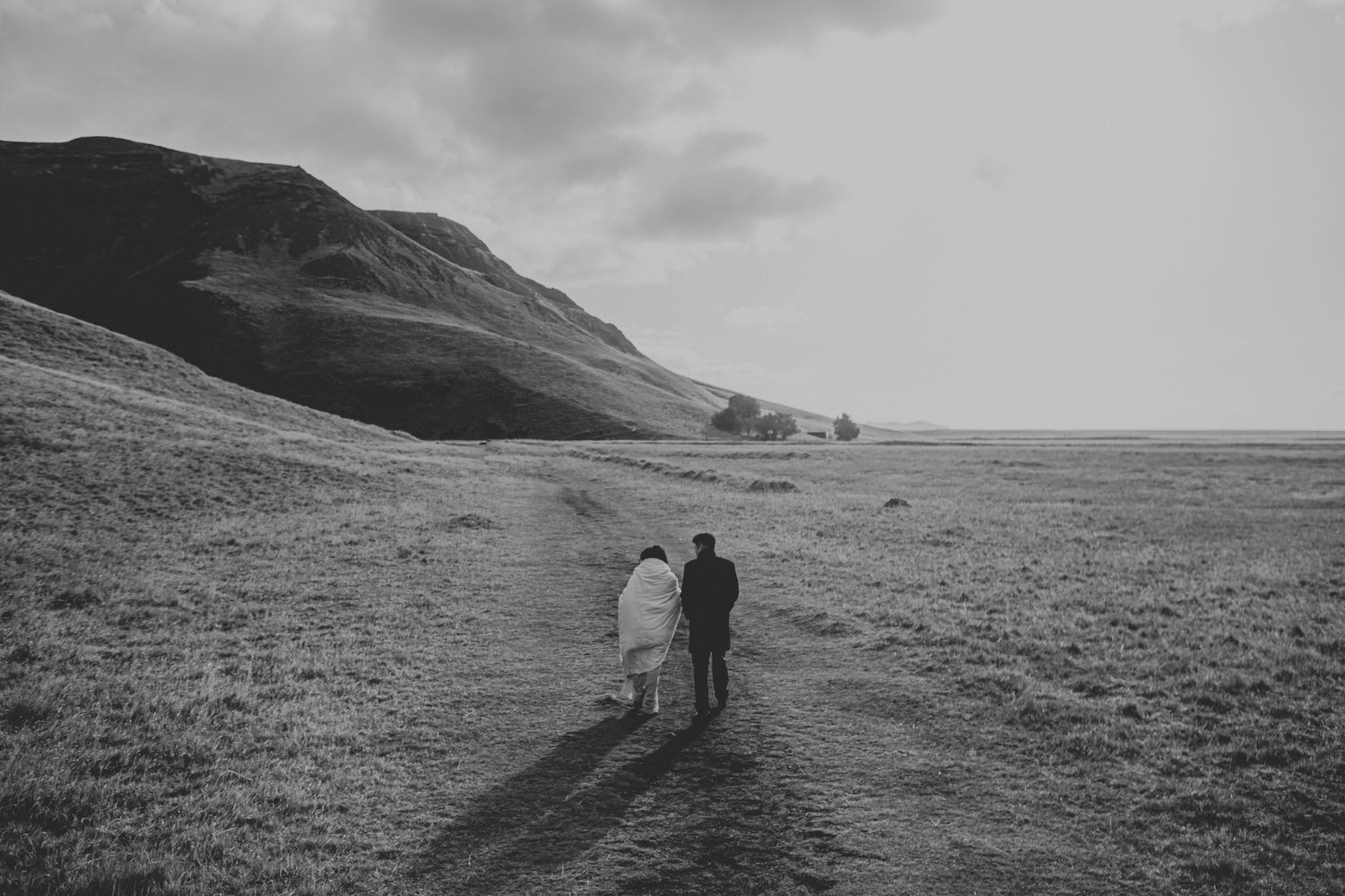Two figures walking along a winding path through a dramatic hillside landscape in black and white.