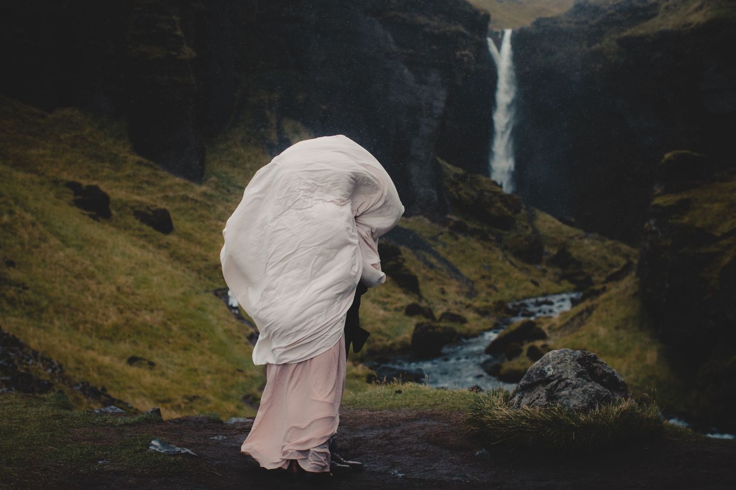 Figure in flowing white dress stands before dramatic waterfall and mountain landscape.