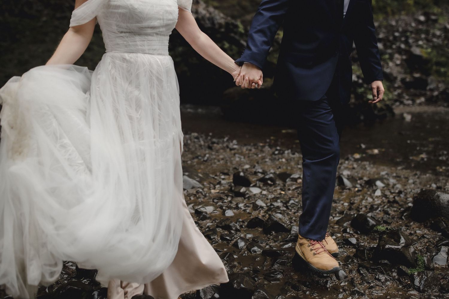 Bride and groom holding hands walking on rocky terrain, bride in flowing white gown, groom in navy suit with tan shoes.
