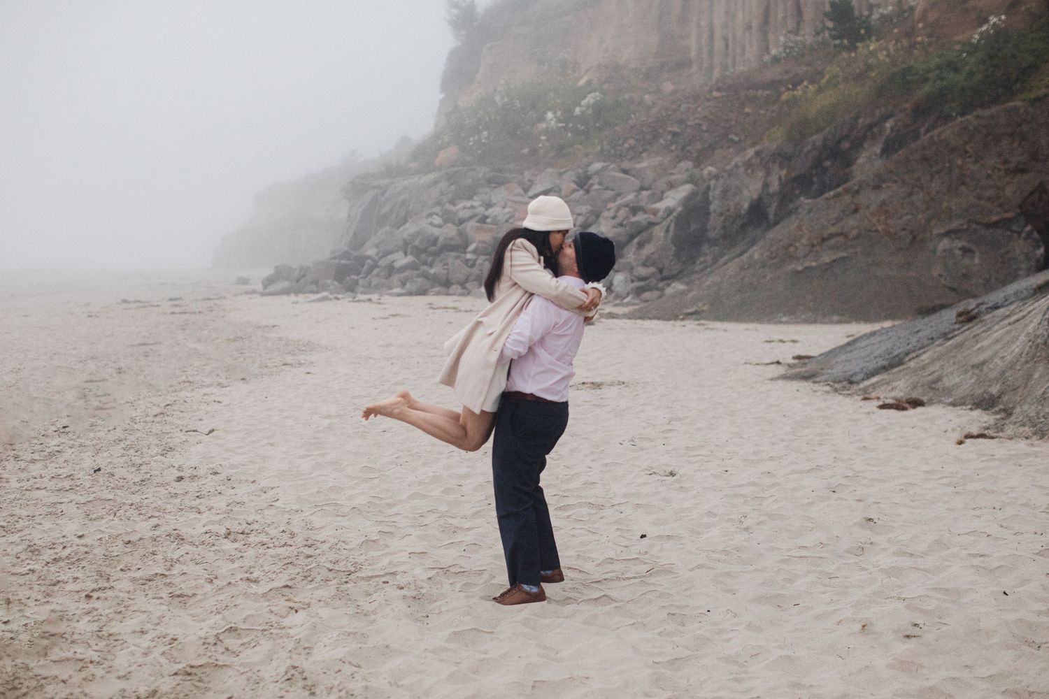 Man lifting woman in a joyful embrace on a foggy beach coastline.