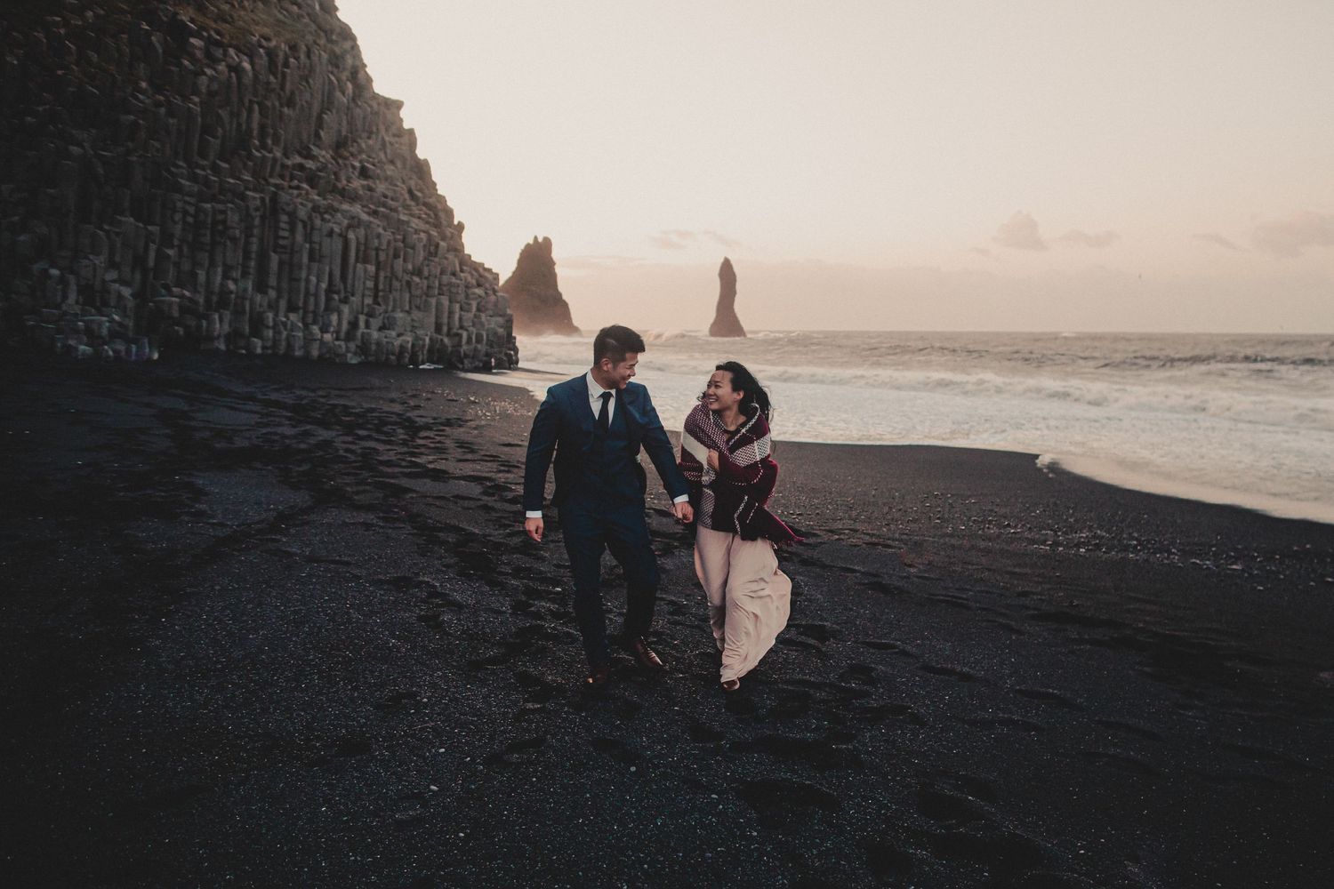A couple walks along a black sand beach with dramatic coastal cliffs and sea stacks at sunset.