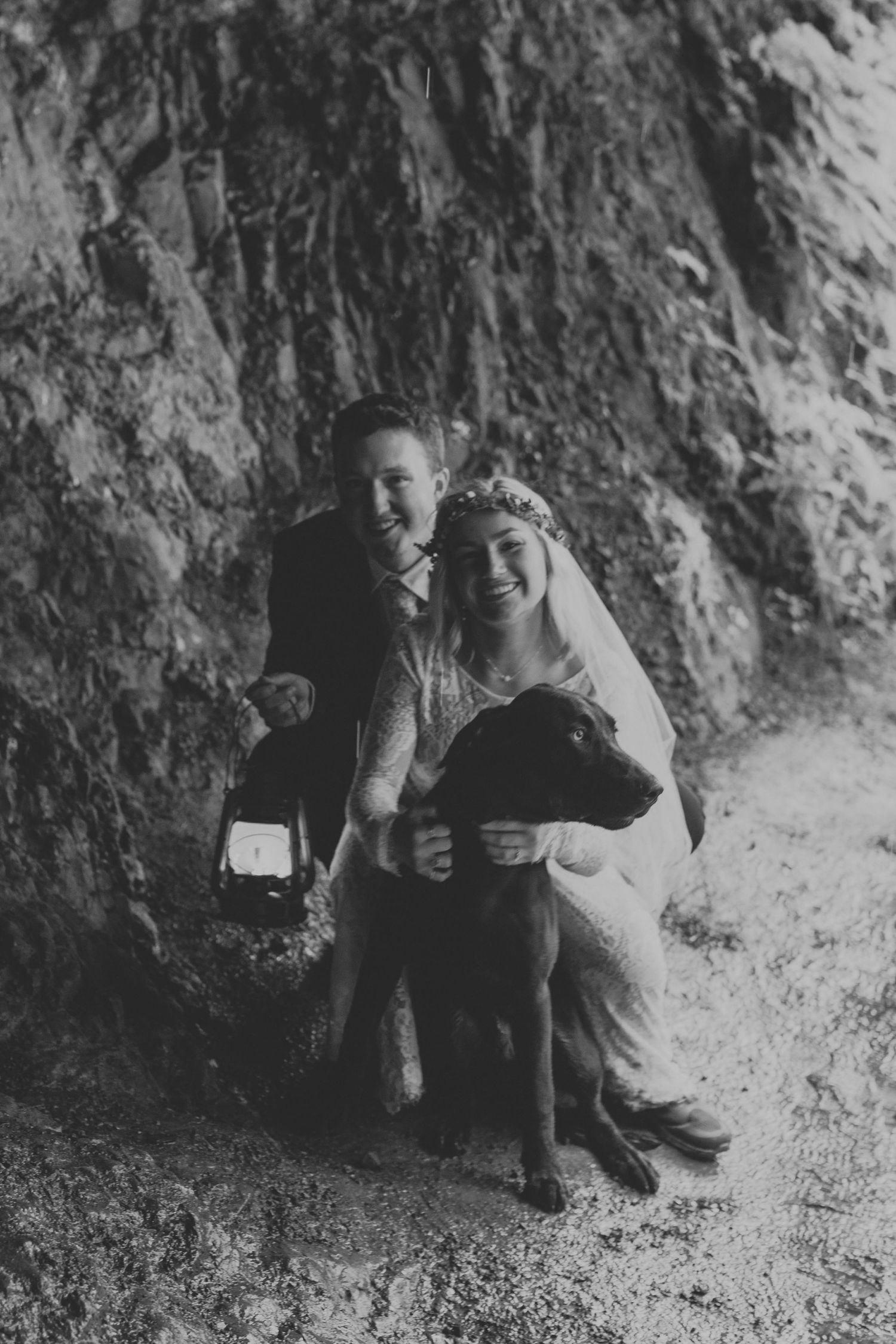 Black and white portrait of newlyweds with their dog against a rocky cliff face.