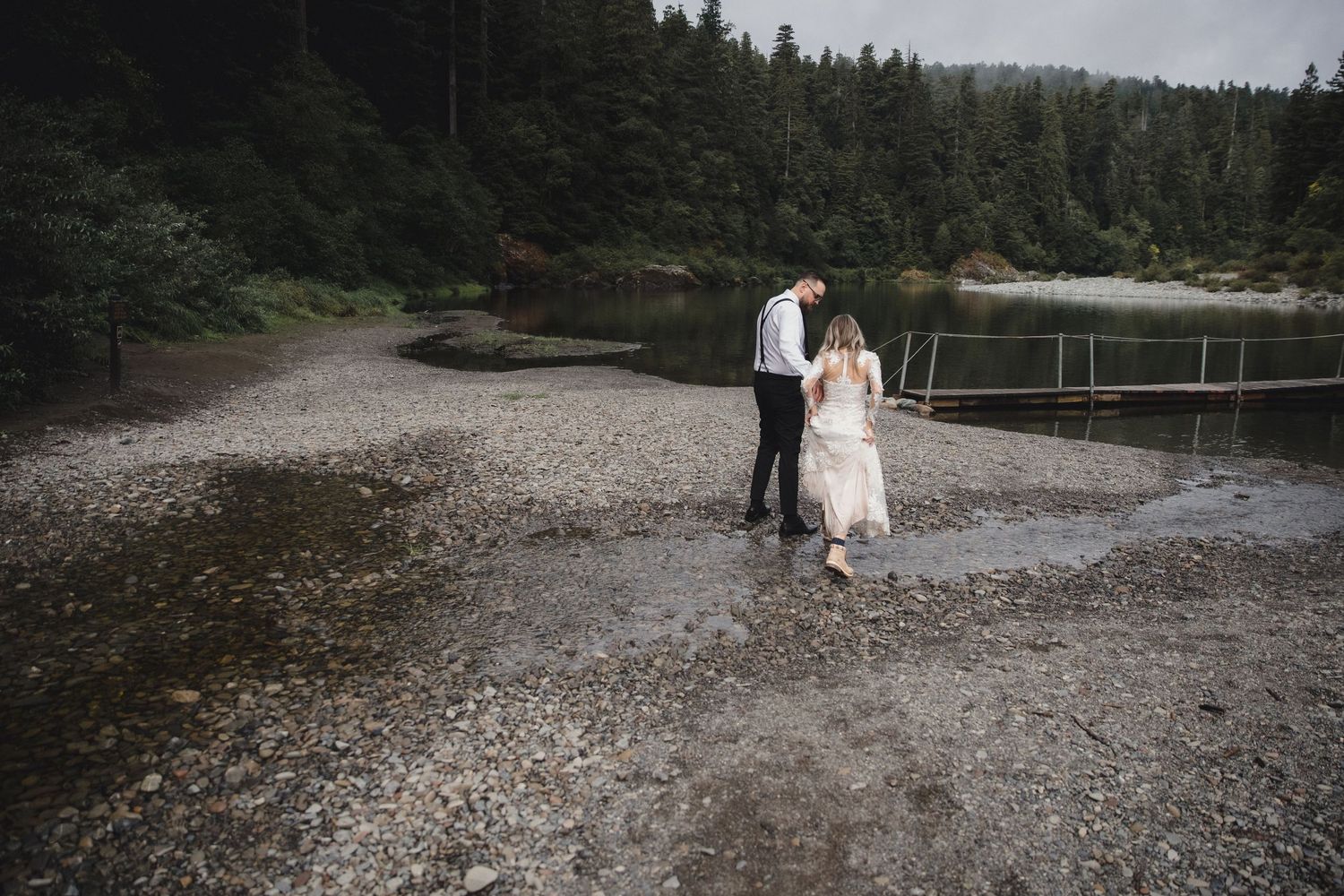 Bride in white gown and groom walk along a rocky shoreline beside a calm lake surrounded by evergreen forest.