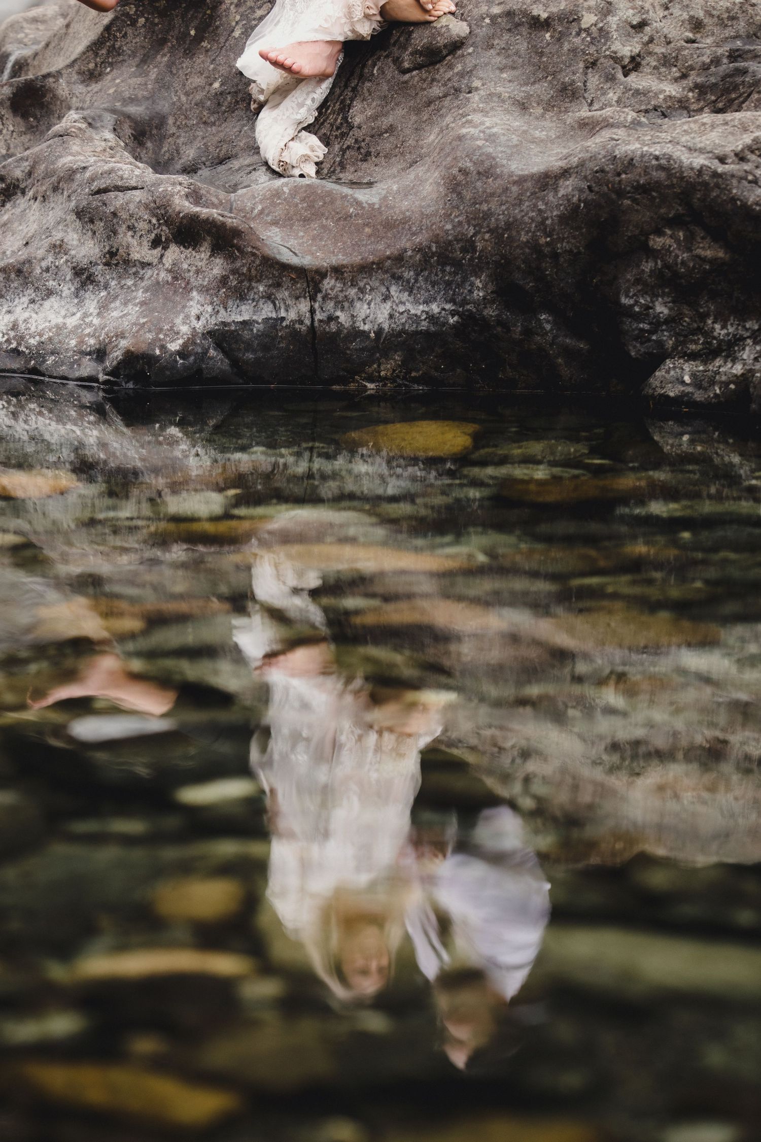 A bride's reflection shimmers in a rocky mountain stream during an outdoor wedding photo session.
