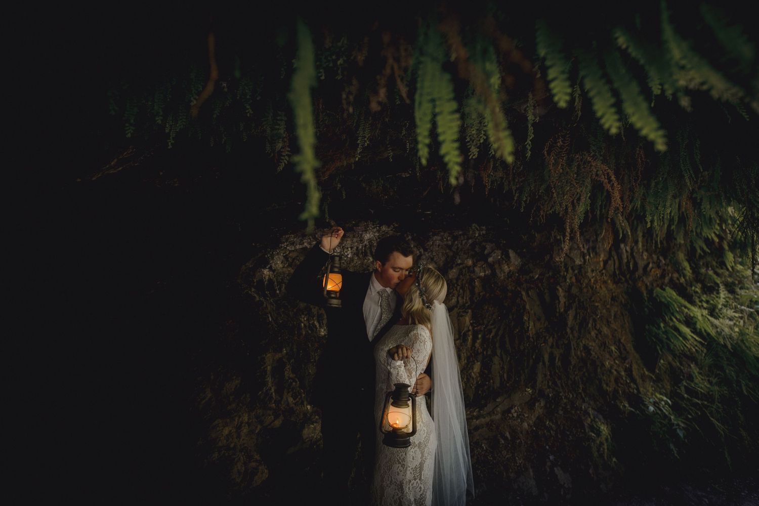 Wedding couple stands under hanging ferns with lantern light creating a dramatic nighttime atmosphere.
