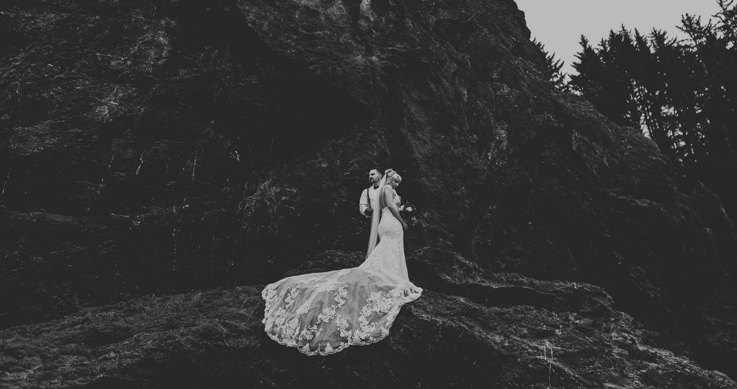Dramatic black and white wedding photo on rocky coastal cliffs with flowing dress.