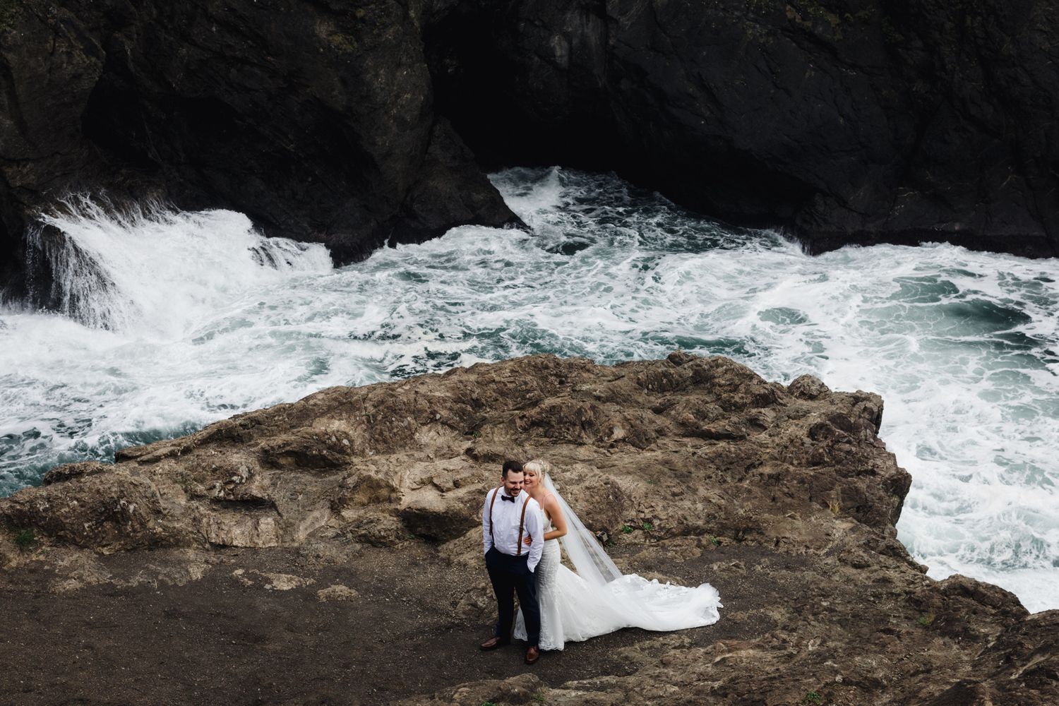 Wedding portrait on rocky coastline with powerful ocean waves crashing below.