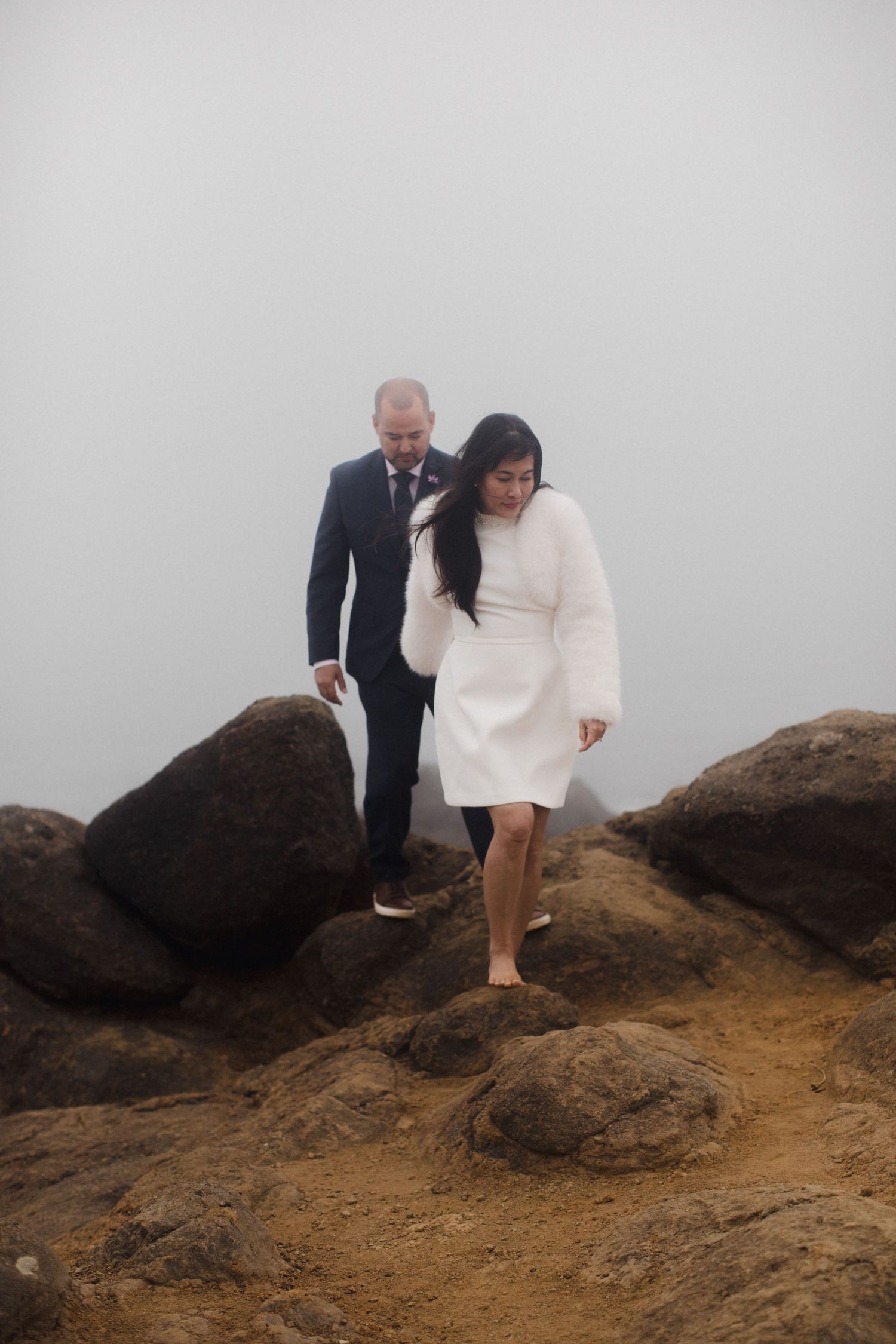 Couple walking on foggy rocky hillside, man in suit and woman in white dress.