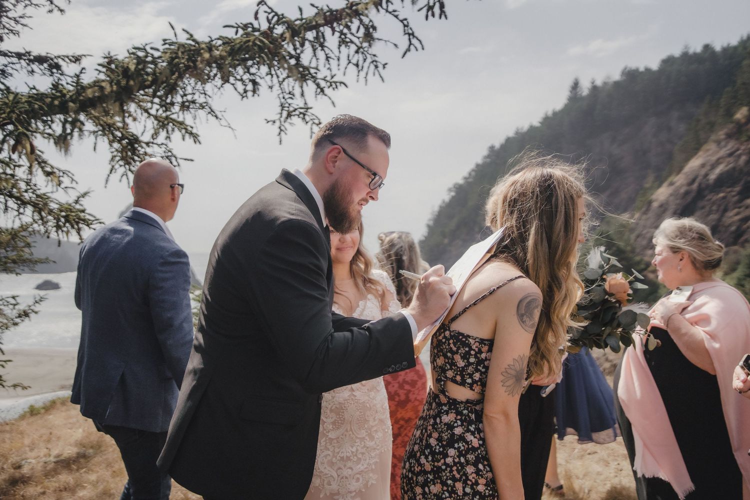 A groom tenderly adjusts his bride's veil during an outdoor coastal wedding ceremony with guests.