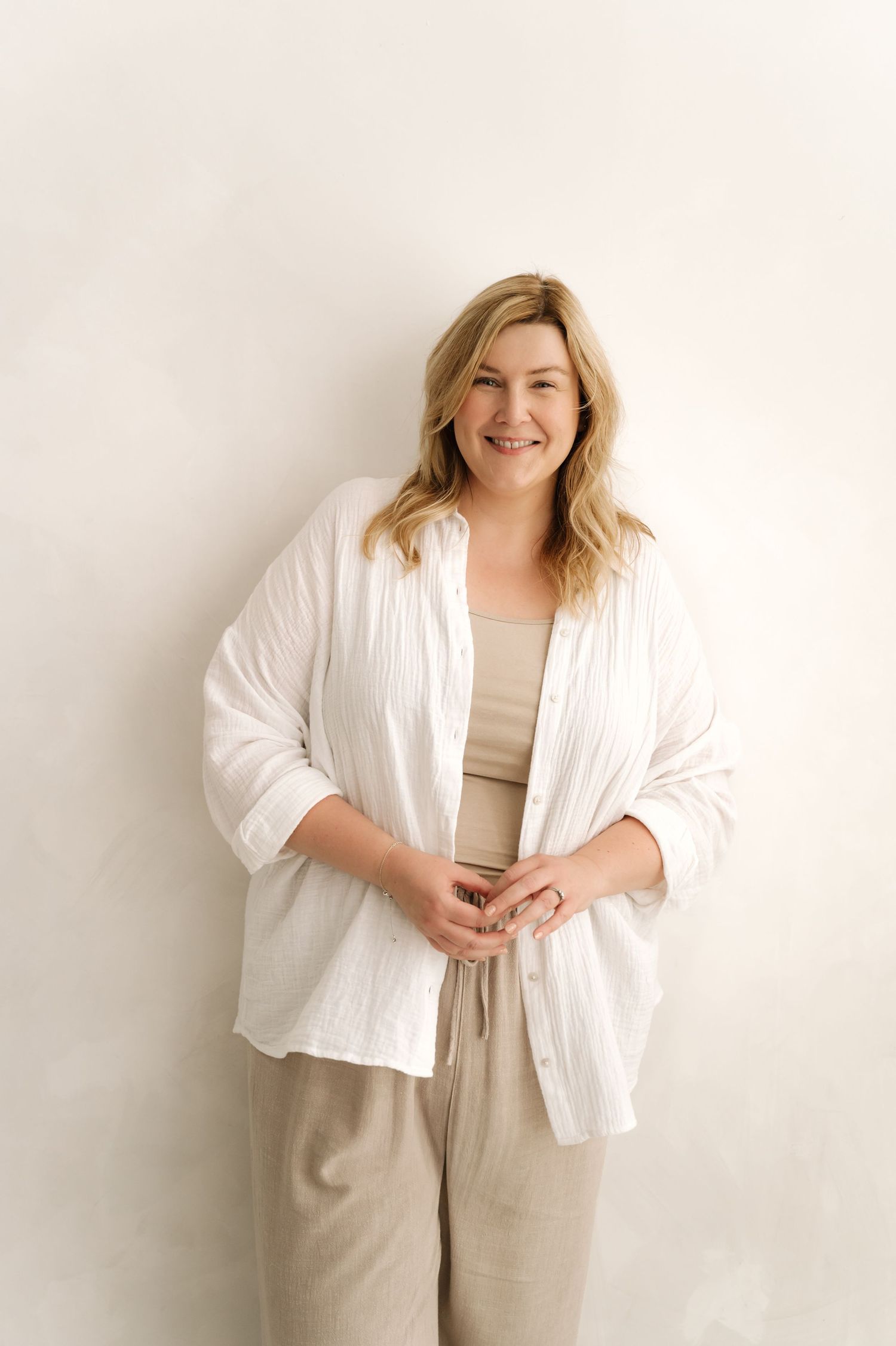 Smiling blonde woman in white linen shirt and beige pants posing against a light background.