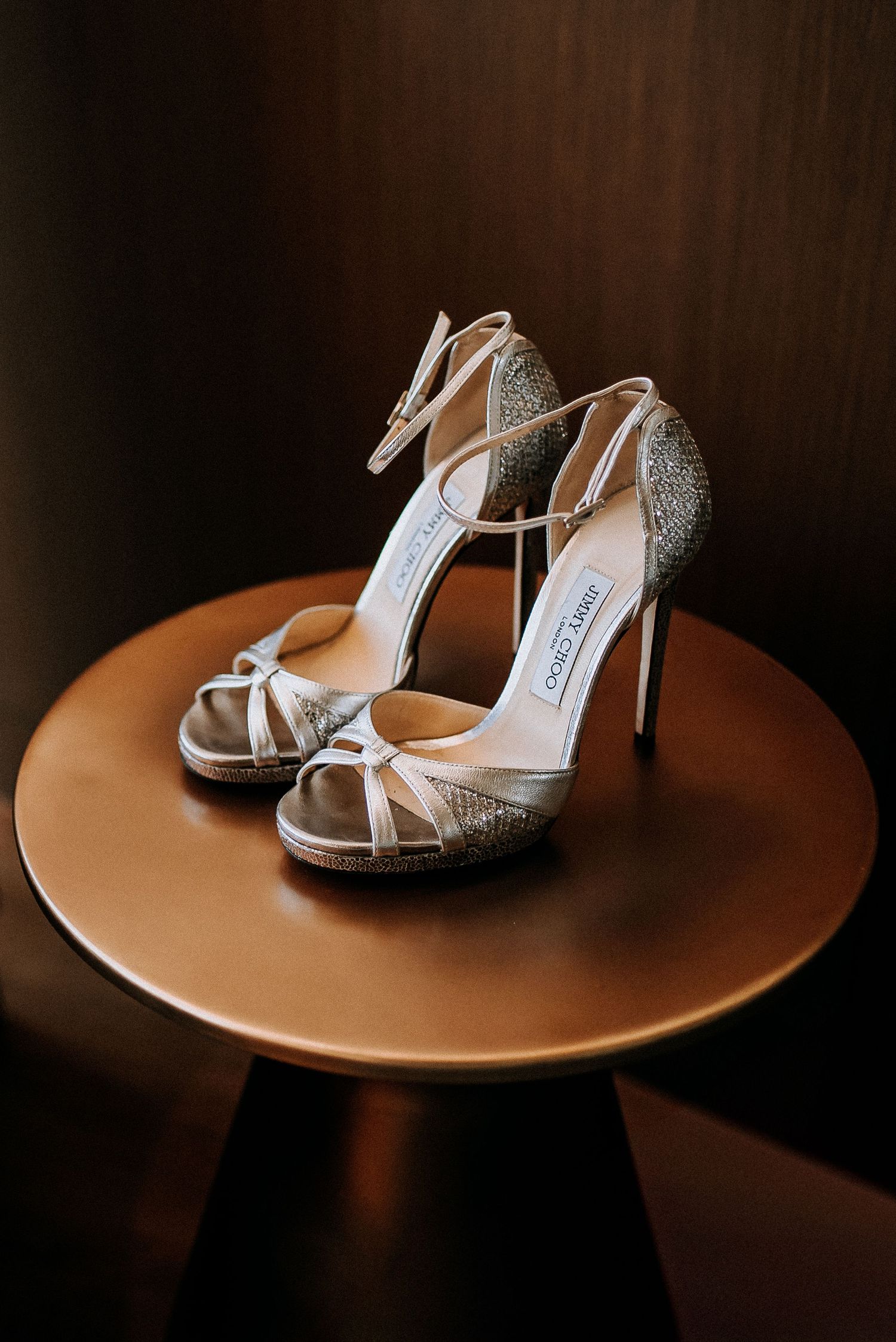 Elegant Jimmy Choo silver glitter strappy high heel sandals displayed on a round wooden table.