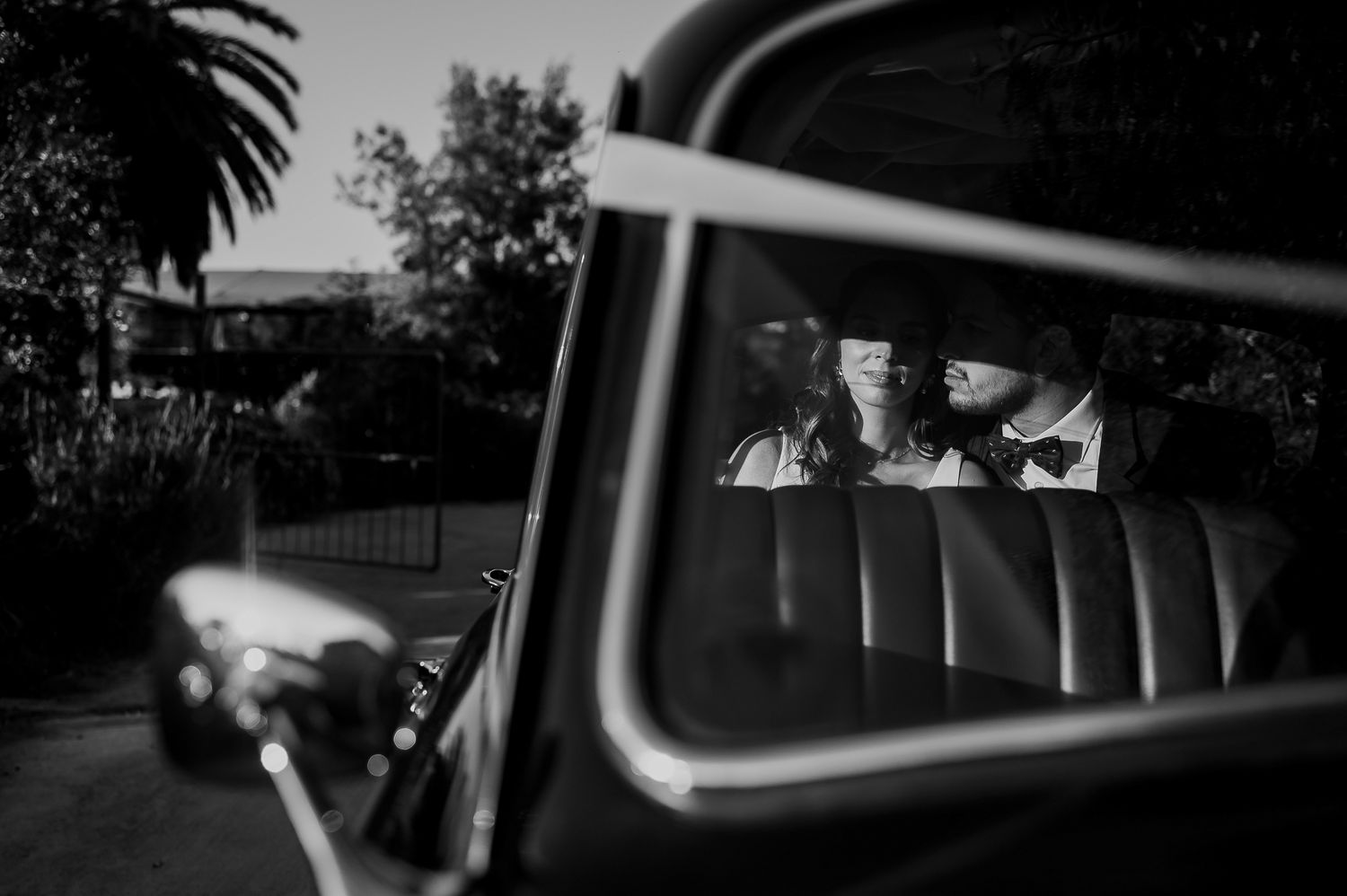 Black and white photo of a couple in formal wear reflected in the window of a vintage classic car.