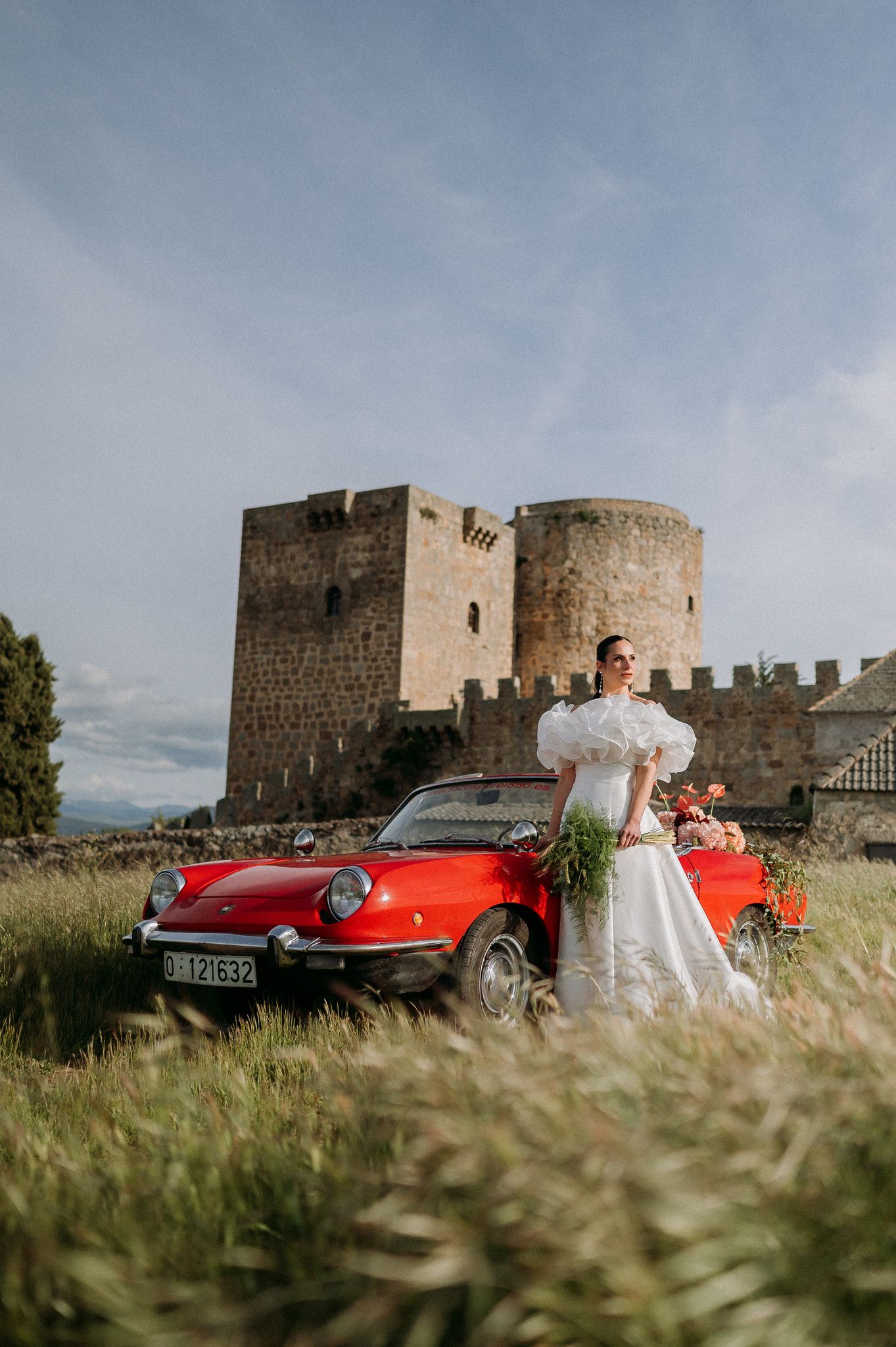 Bride in white gown poses by vintage red convertible car in front of ancient stone castle tower in green field.