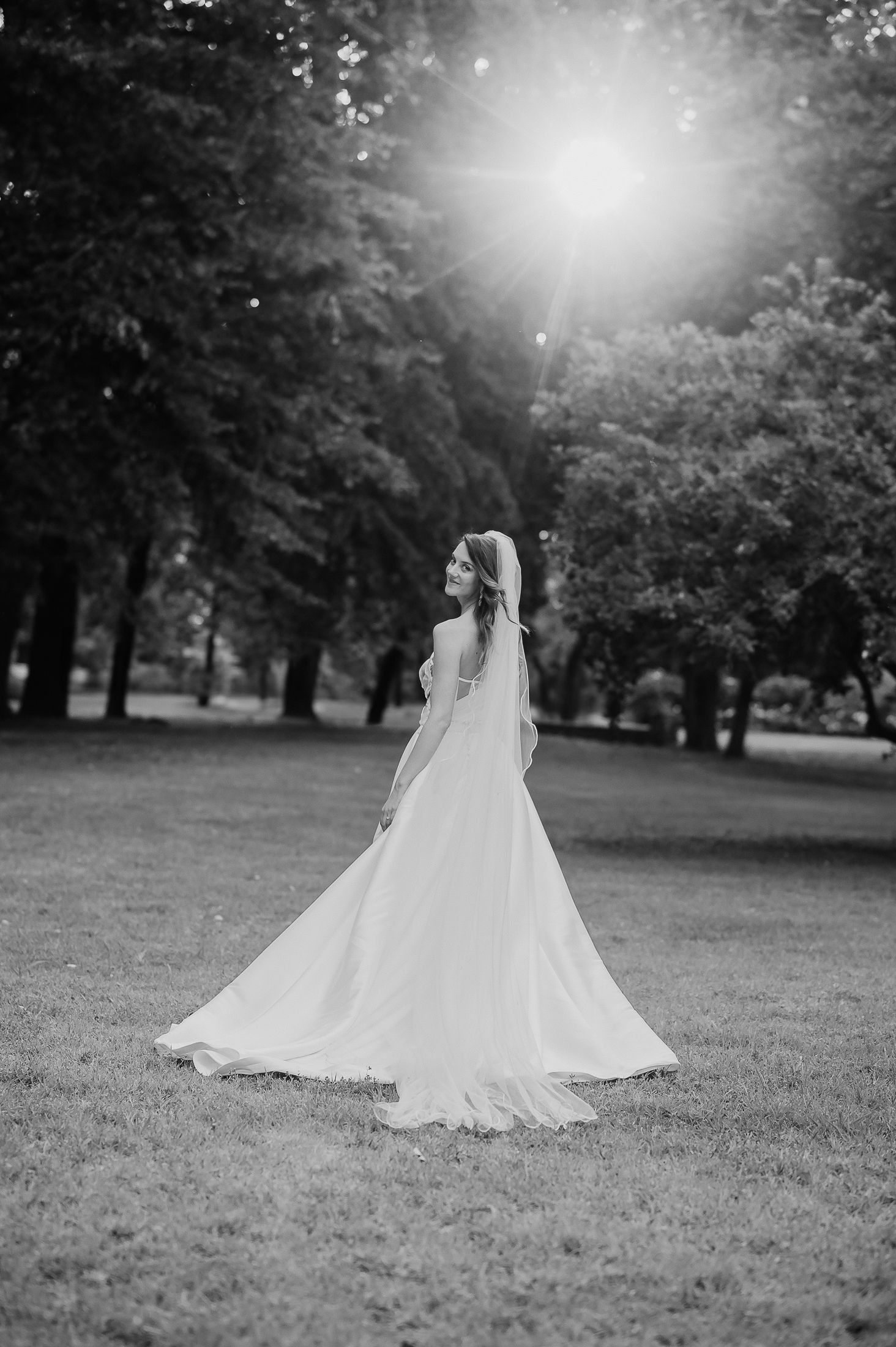 Black and white photo of a bride in a flowing white gown standing in a sunlit park with trees in the background.