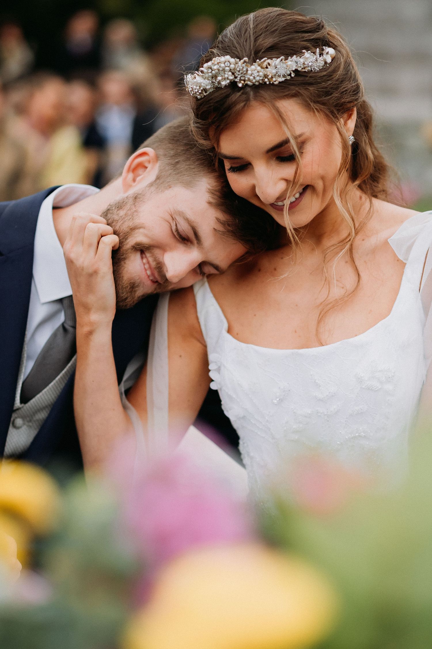 A joyful bride in a white gown and floral crown leans close to a smiling groom in a navy suit at their wedding.