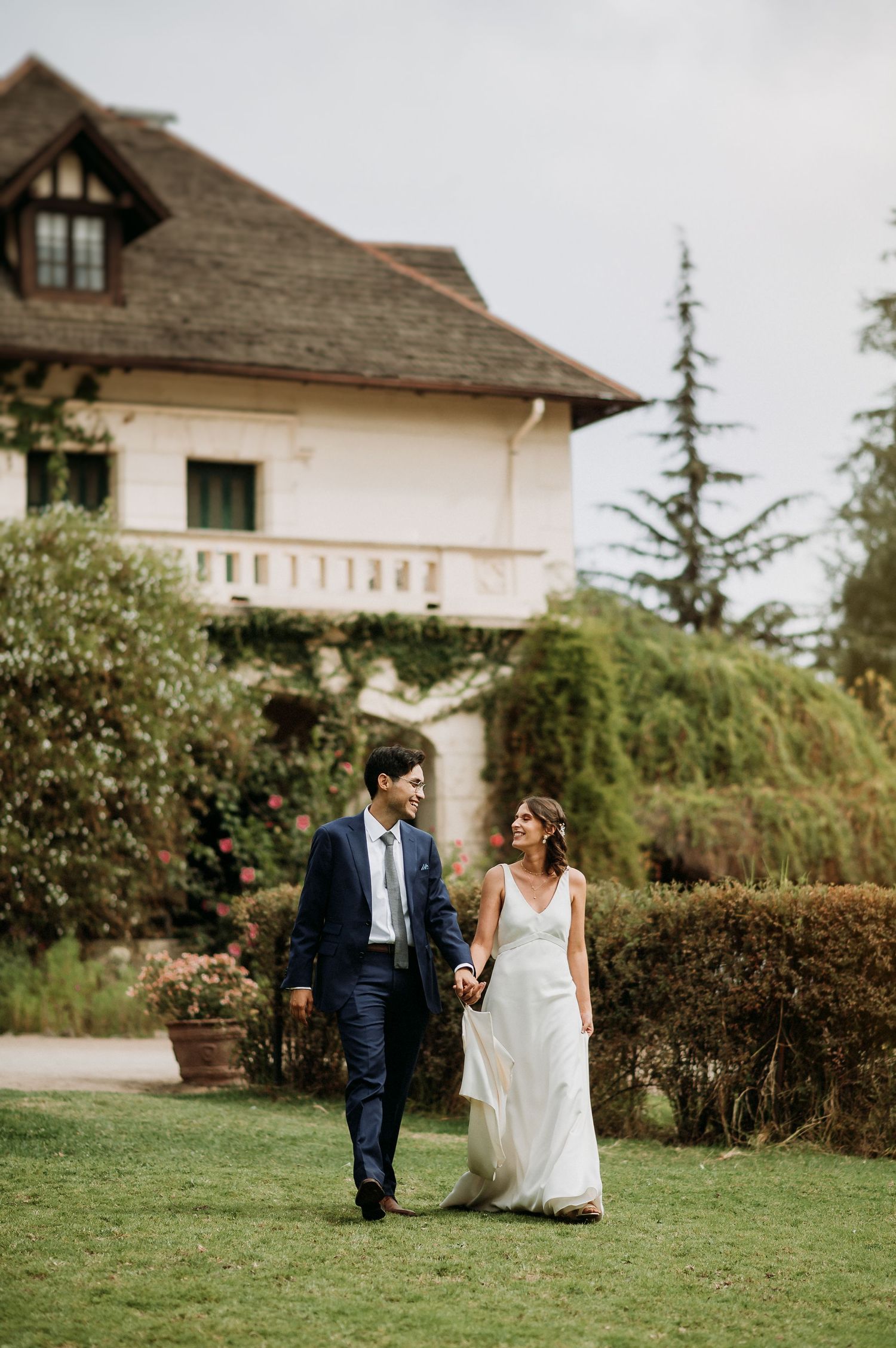 Bride and groom walk hand in hand on a lawn in front of an elegant ivy-covered estate.