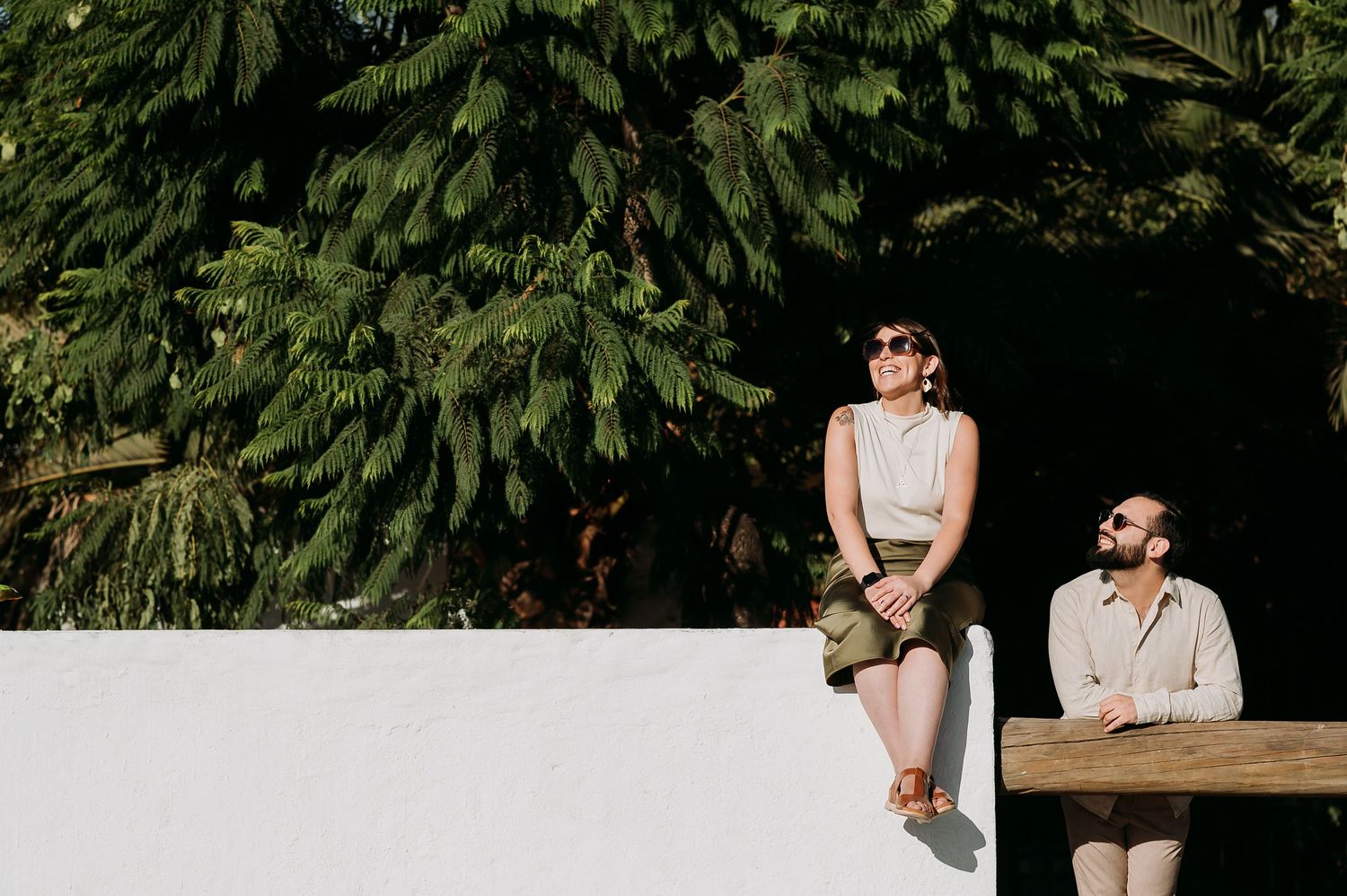 Couple relaxing outdoors near white wall with lush green trees, woman sitting and man standing nearby.