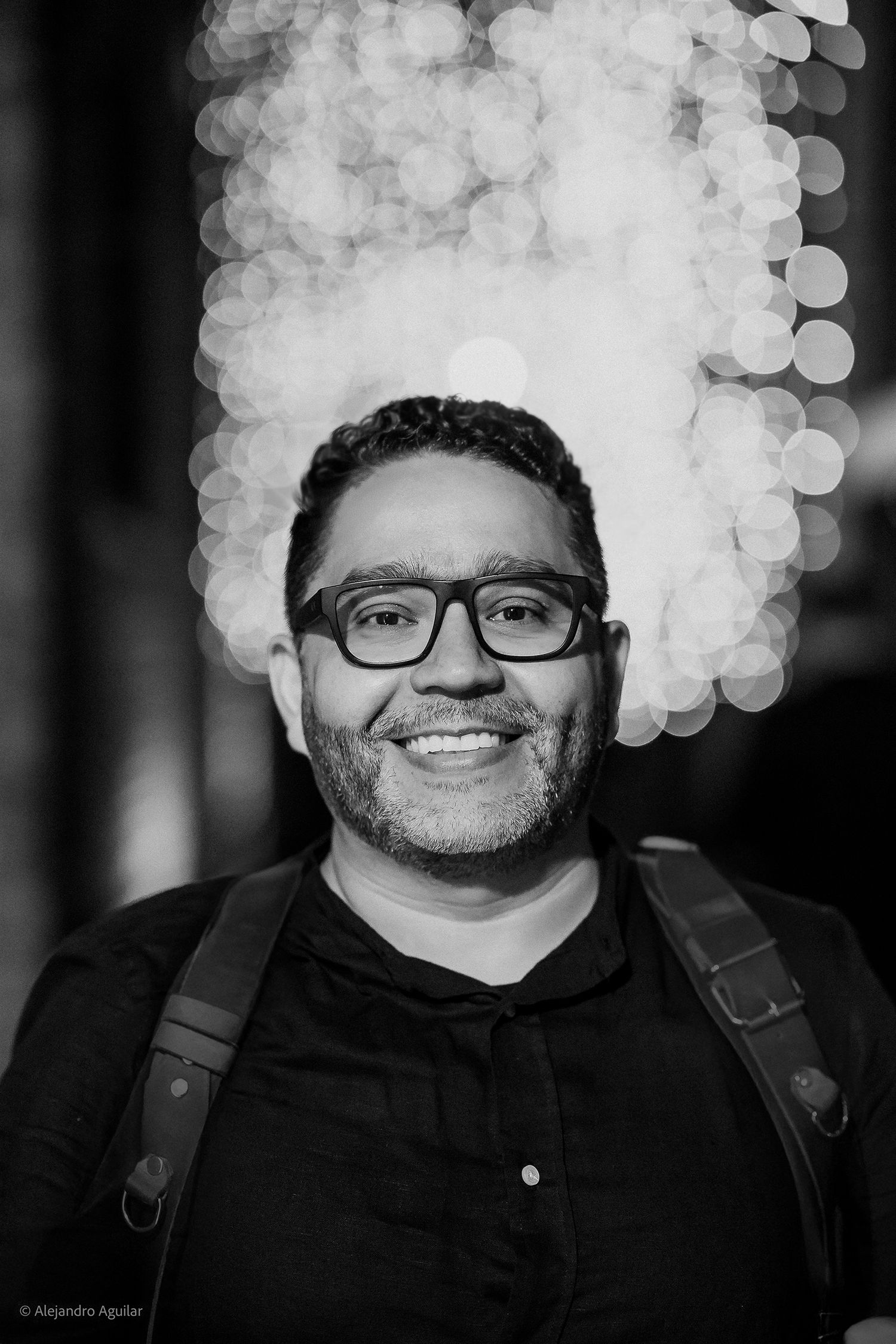 Black and white portrait of smiling man with glasses and backpack against bokeh light background.