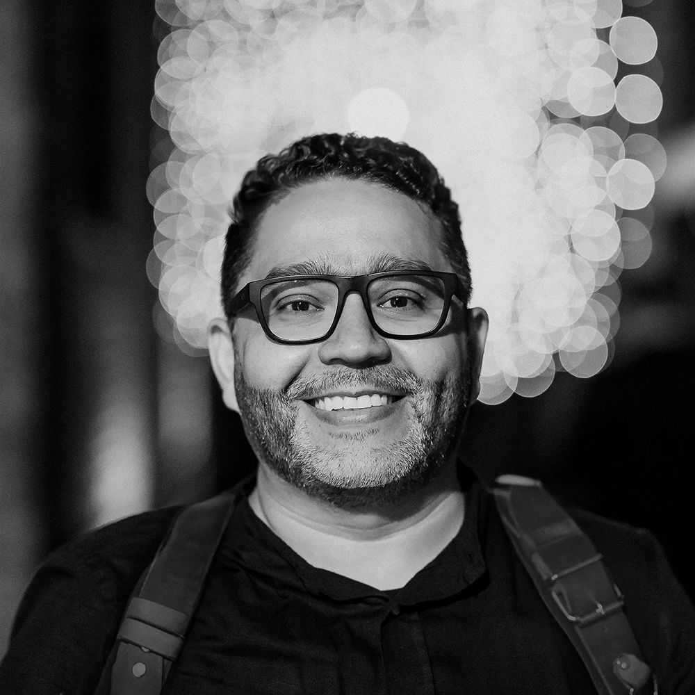 Black and white portrait of a smiling man wearing glasses and a backpack with bokeh lights in background.