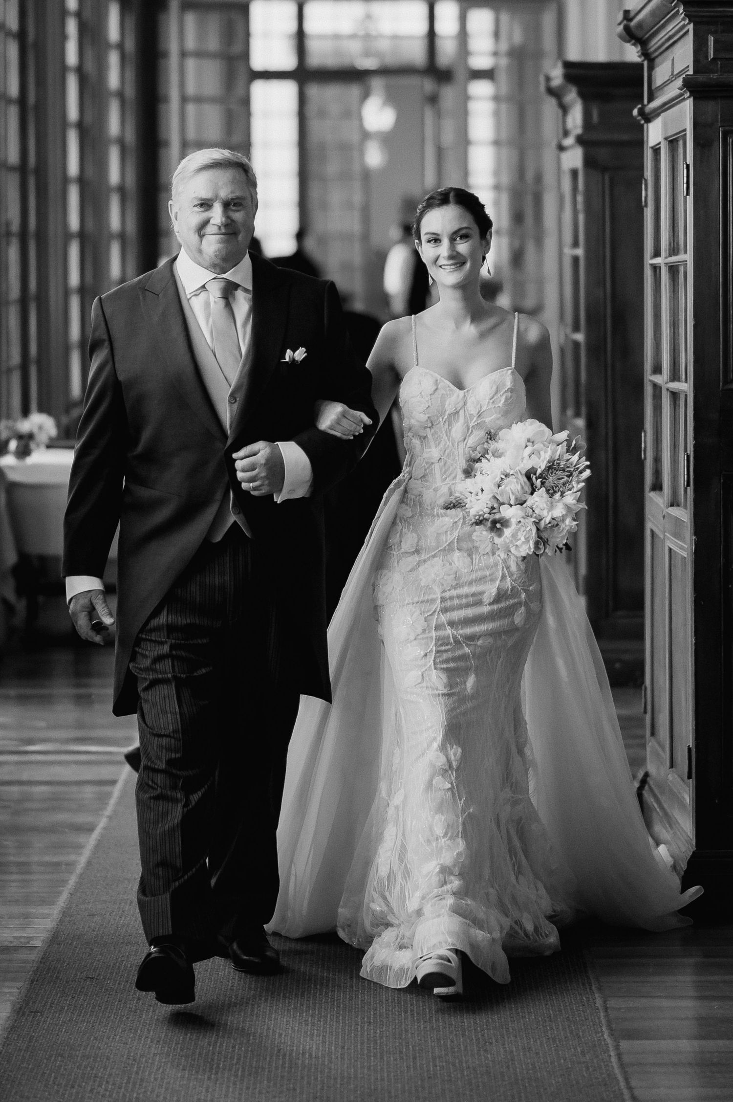 Black and white photo of a bride in a lace gown walking down the aisle with her father in a suit.